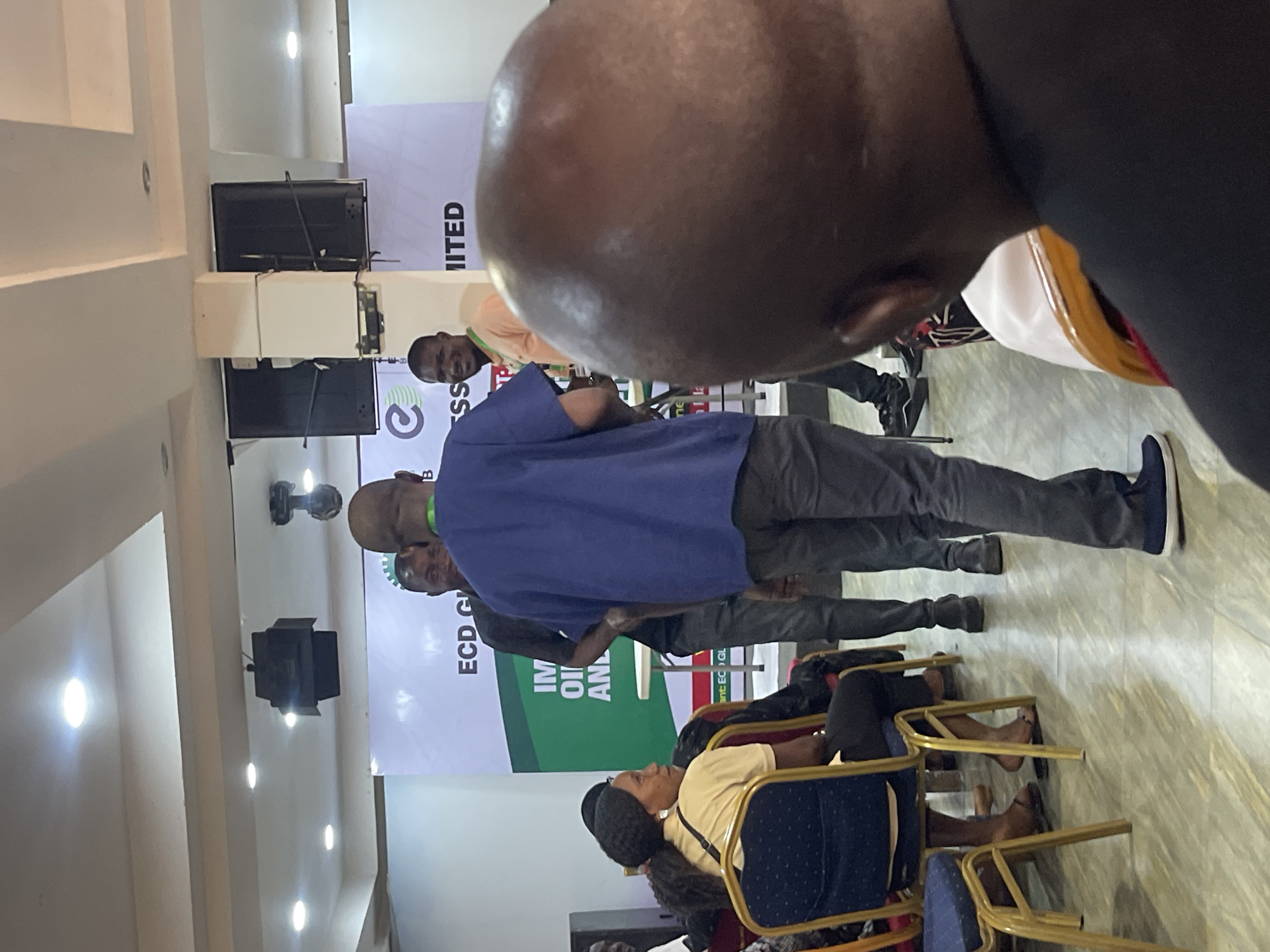 The image shows an indoor scene in what appears to be a conference or meeting room in Galadimawa Village, Nigeria. People are seen standing and sitting. A man in a blue shirt stands facing toward a banner; the banner reads "ECD G," and includes the text "OIL AND." A woman in a yellow shirt sits on a chair. The room is lit by ceiling lights. There are additional people in the background, along with a banner with partial text "LIMITED". Audio visual equipment and speakers can be seen mounted on the wall.