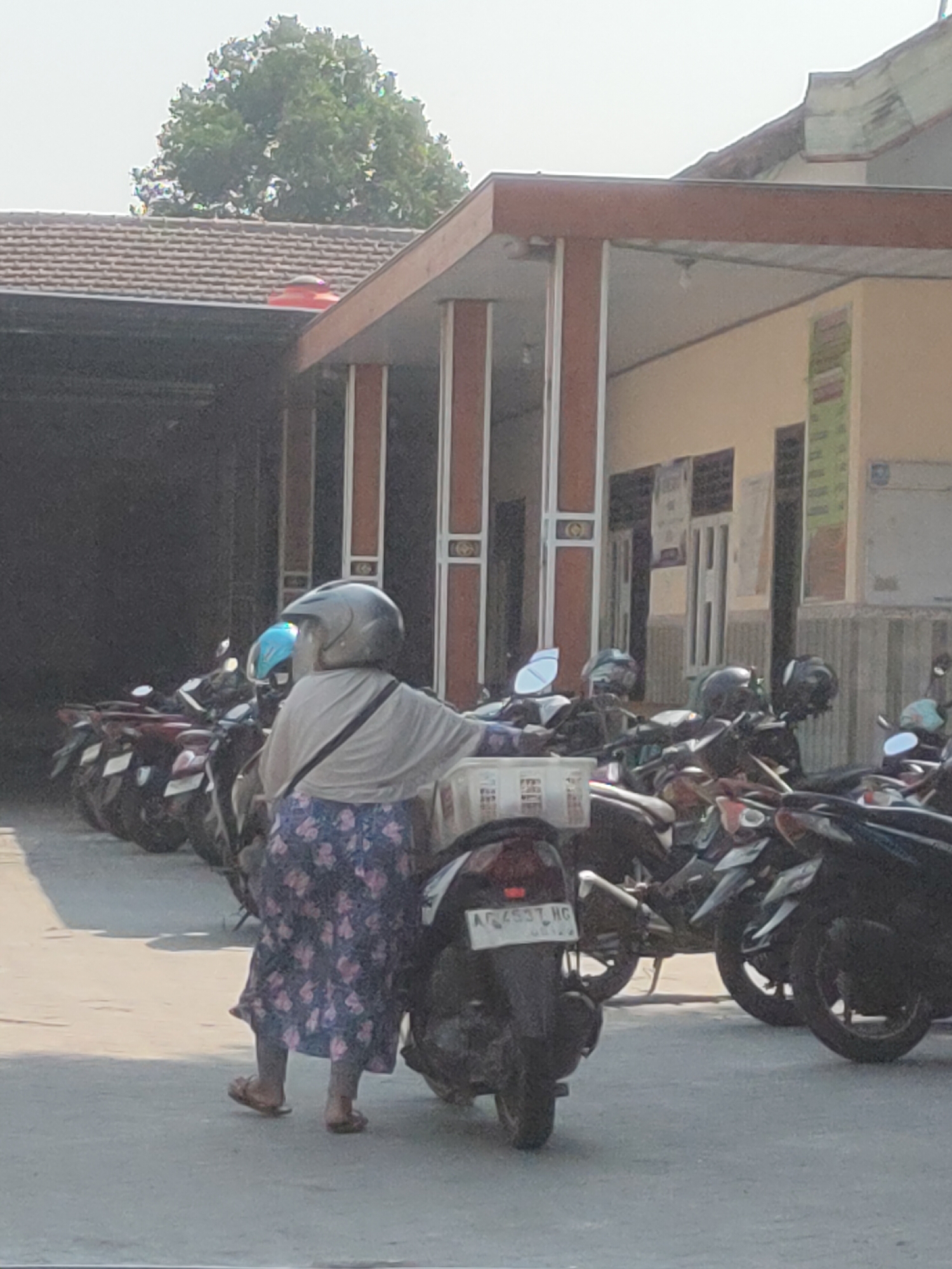 The image captures a scene in front of a building with a tiled roof and terracotta-colored pillars, likely in Ngasem, Indonesia. In the foreground, a person wearing a grey helmet, a light-colored shawl, and a floral-patterned skirt is standing next to a dark-colored motorcycle. They are holding a white plastic basket. The motorcycle has a license plate with the characters "AQ4537 HG". Behind this person, several other motorcycles are parked in rows, some with helmets on their handlebars. The background shows a building with windows and posters on the wall, and a large green tree visible above the roofline. The overall lighting suggests it is a sunny day.
