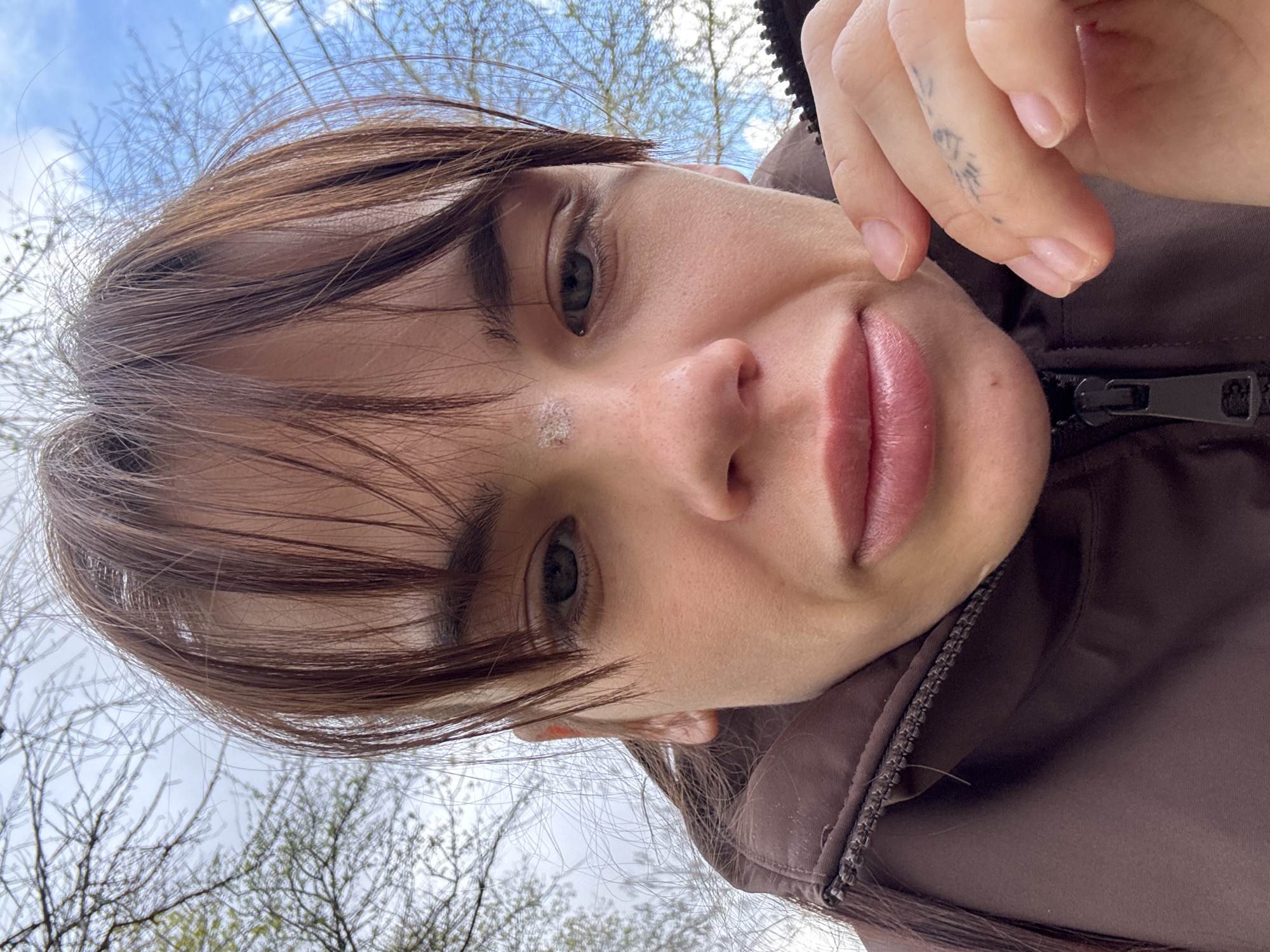 A close-up selfie taken from a low angle shows a woman with brown hair and fair skin. She has a small tattoo on her finger. She is wearing a brown jacket with a visible zipper. In the background, out of focus, are trees and a blue sky with clouds. There is no information to place the location in Rostov-on-Don.
