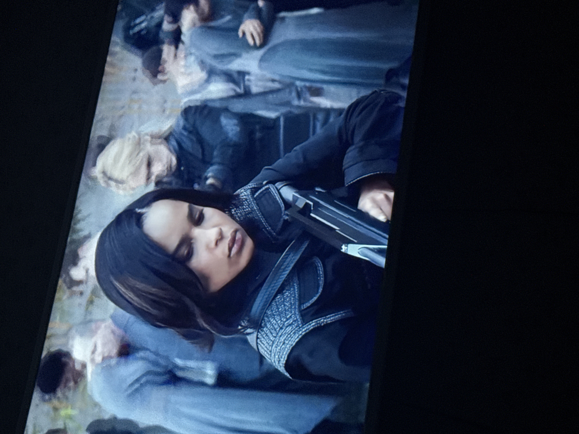 A medium shot of a scene from a movie or television show. In the foreground, a woman with dark hair is lying down, holding a firearm across her body. She is wearing a dark jacket and a gray, woven vest-like garment. Behind her, other people are lying on the ground, also seemingly unconscious. The background is blurry and indistinct, but it appears to be an outdoor setting. The image seems to be taken from a screen or monitor. There is no information to describe the location in the city of Mytishchi, Russia.