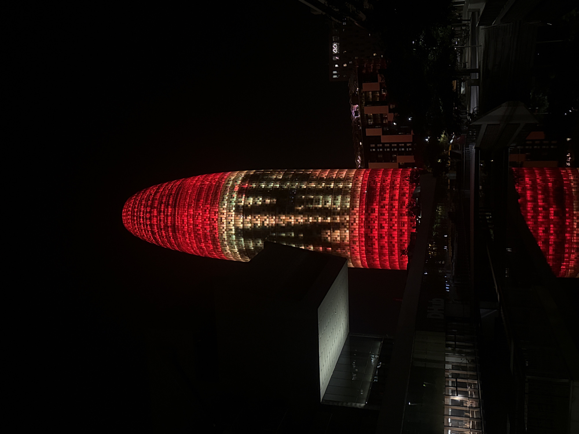 This is a nighttime, eye-level, medium shot of the Torre Glòries skyscraper in Barcelona, Spain. The building is illuminated with red and yellow lights, creating a patterned effect on its facade. To the left of the skyscraper, a modern, dark-colored building with a cantilevered section is visible. In the foreground, there is a body of water reflecting the lights of the buildings. The scene is dark, with the exception of the illuminated structures and some scattered lights from other buildings in the background. The overall impression is urban and modern.