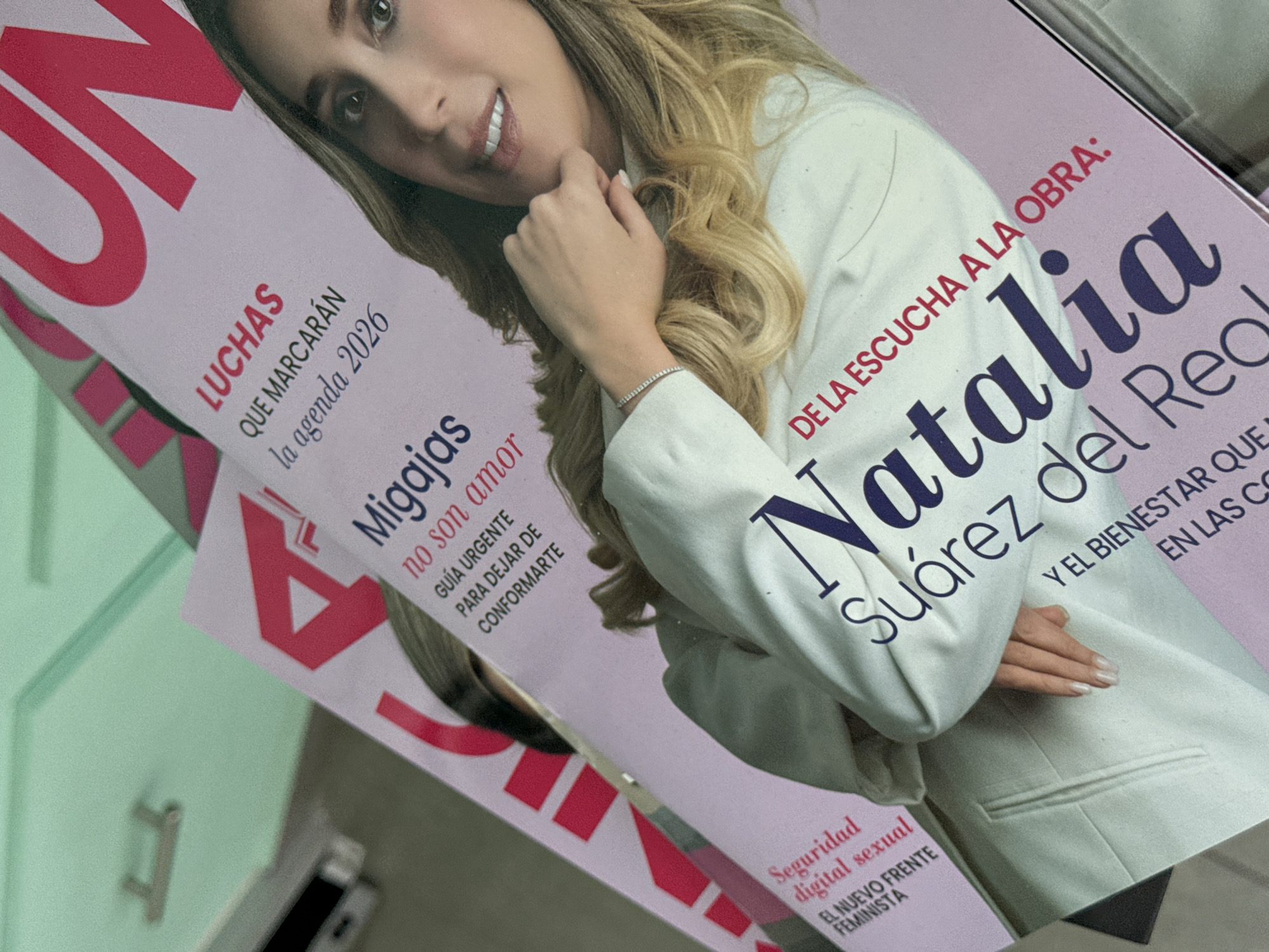 This media file presents a close-up, angled view of a magazine cover, prominently featuring a smiling woman identified by the cover text as Natalia Suárez del Real. The woman has long blonde hair, light makeup, and is wearing a white blazer, with a thin silver bracelet on her wrist. Her hand is gently positioned near her chin, conveying a composed demeanor. The magazine cover is primarily light pink, adorned with red and dark blue text in Spanish.
Visible headlines on the cover include: "DE LA ESCUCHA A LA OBRA:" (From listening to action:), the main subject's name "Natalia Suárez del Real," and "Y EL BIENESTAR QUE EN LAS CO" (And the well-being that in the co...). On the left side of the cover, other article titles are "LUCHAS QUE MARCARÁN la agenda 2026" (Struggles that will mark the 2026 agenda), "Migajas no son amor: GUÍA URGENTE PARA DEJAR DE CONFORMARTE" (Crumbs are not love: Urgent guide to stop settling), and "Seguridad digital sexual EL NUEVO FRENTE FEMINISTA" (Digital sexual security: The new feminist front). The background is an indistinct, soft light green, suggesting an indoor setting. The photograph of the magazine is taken from an elevated, tilted perspective. The overall impression is that of a professional publication with themes of social relevance.