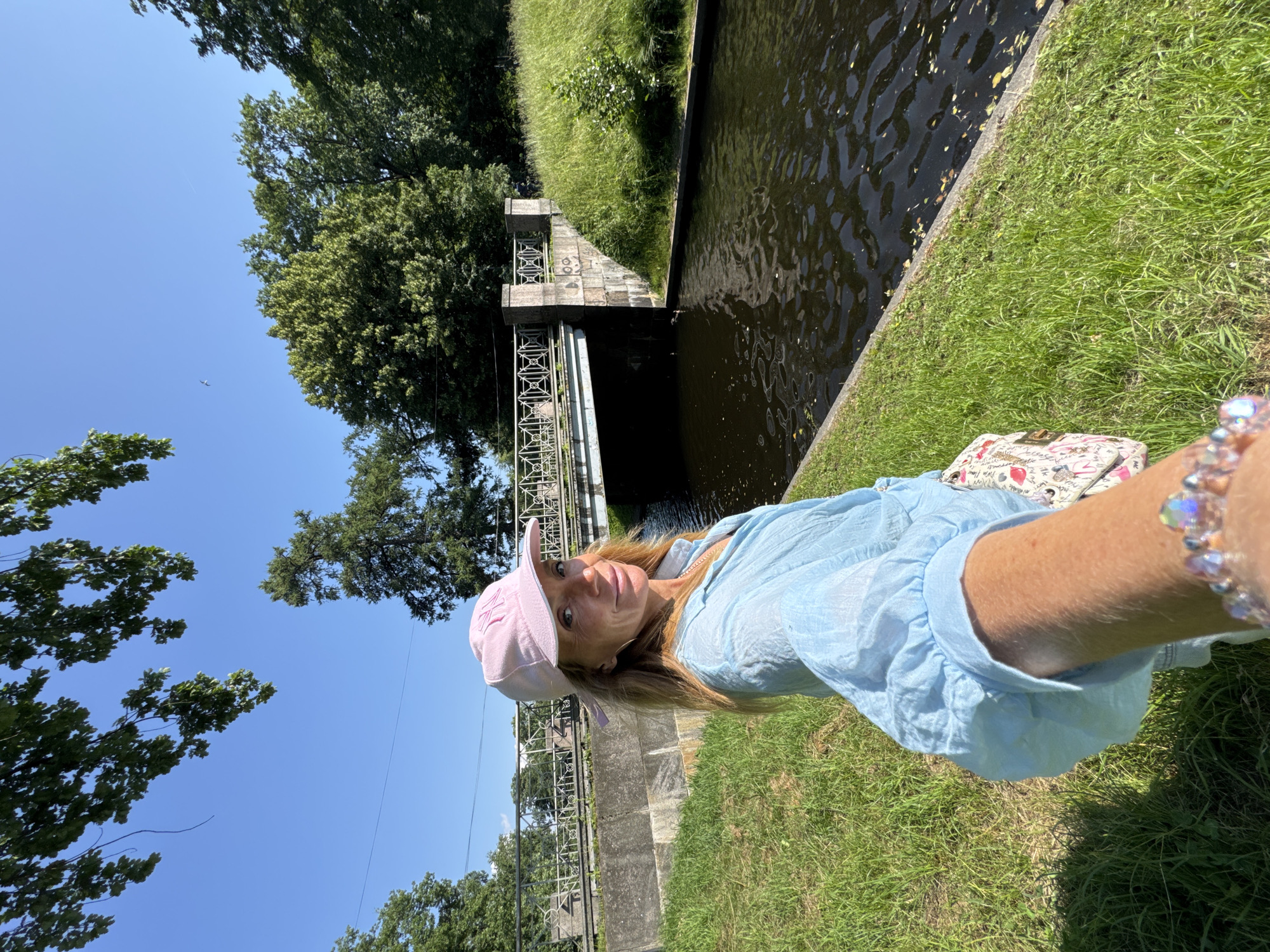 A woman with blonde hair, wearing a light blue shirt and a pink baseball cap, is taking a selfie. She has a bright and engaged expression. The scene is a sunny day outdoors, likely in the afternoon, given the quality of light. The woman is sitting on a grassy bank next to a dark, calm body of water that appears to be a canal or a wide river. In the background, there is a stone structure with decorative metal railings, possibly a bridge or a lock gate, typical of European canal systems. Lush green trees and vegetation surround the water and bank, suggesting a park-like setting or a scenic waterfront area. The woman is also wearing a bracelet with clear, multifaceted beads. A patterned bag with a floral motif is visible near her. The overall mood is relaxed and pleasant, indicative of an enjoyable outdoor excursion. Visible text on the baseball cap appears to be "NY".