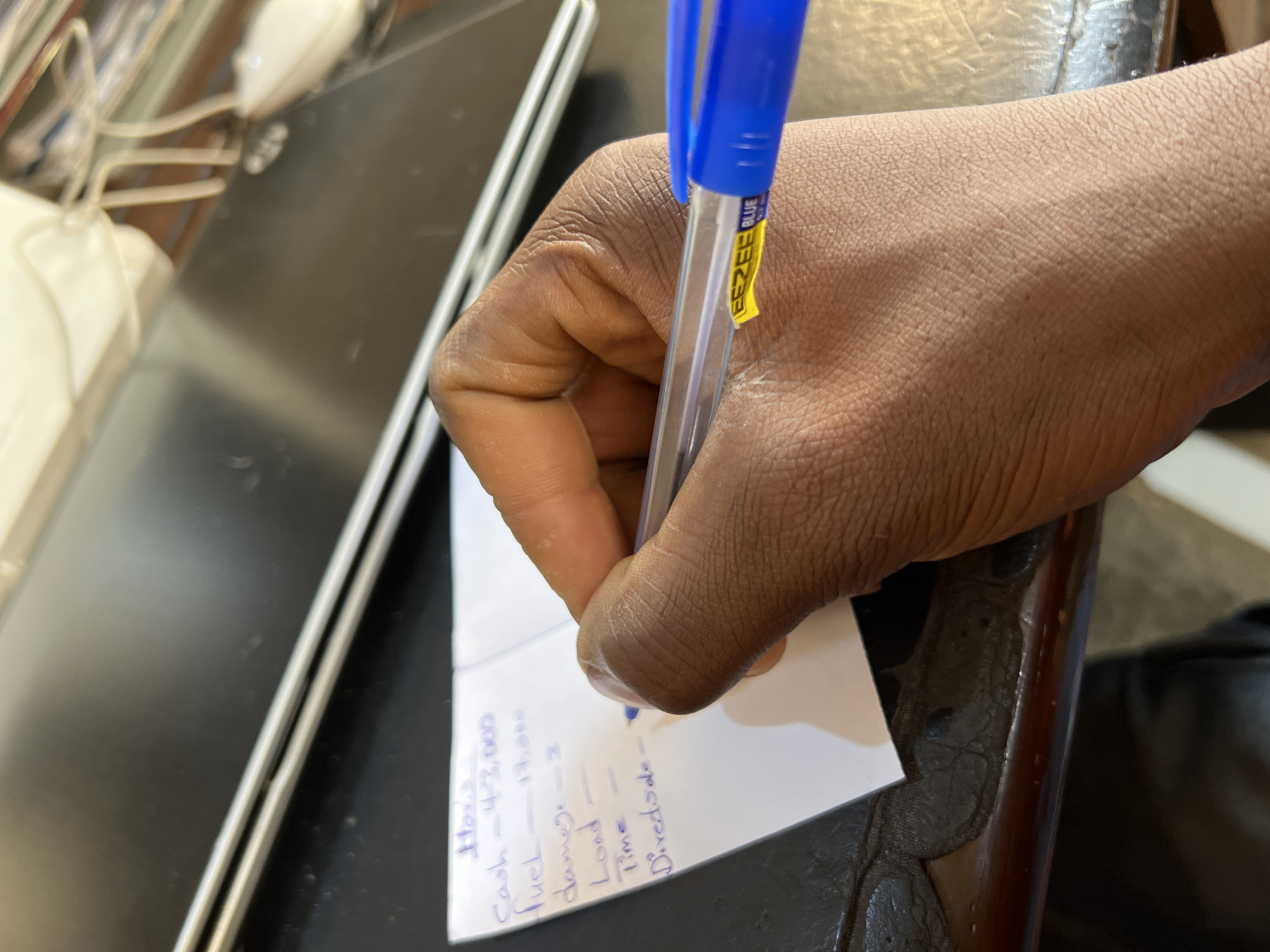 A hand is writing on a piece of paper with a blue pen. The text on the paper includes the words "Hox's", "Cash=43,000", "Fuel=17,000", "damage=2", "Load", "time", and "Dividsde-". The brand of the pen is "EEZEE BLUE". The scene appears to be indoors, on a dark surface with a silver-colored object nearby. The city of Jos, Nigeria is mentioned, but nothing in the image explicitly places the scene there.