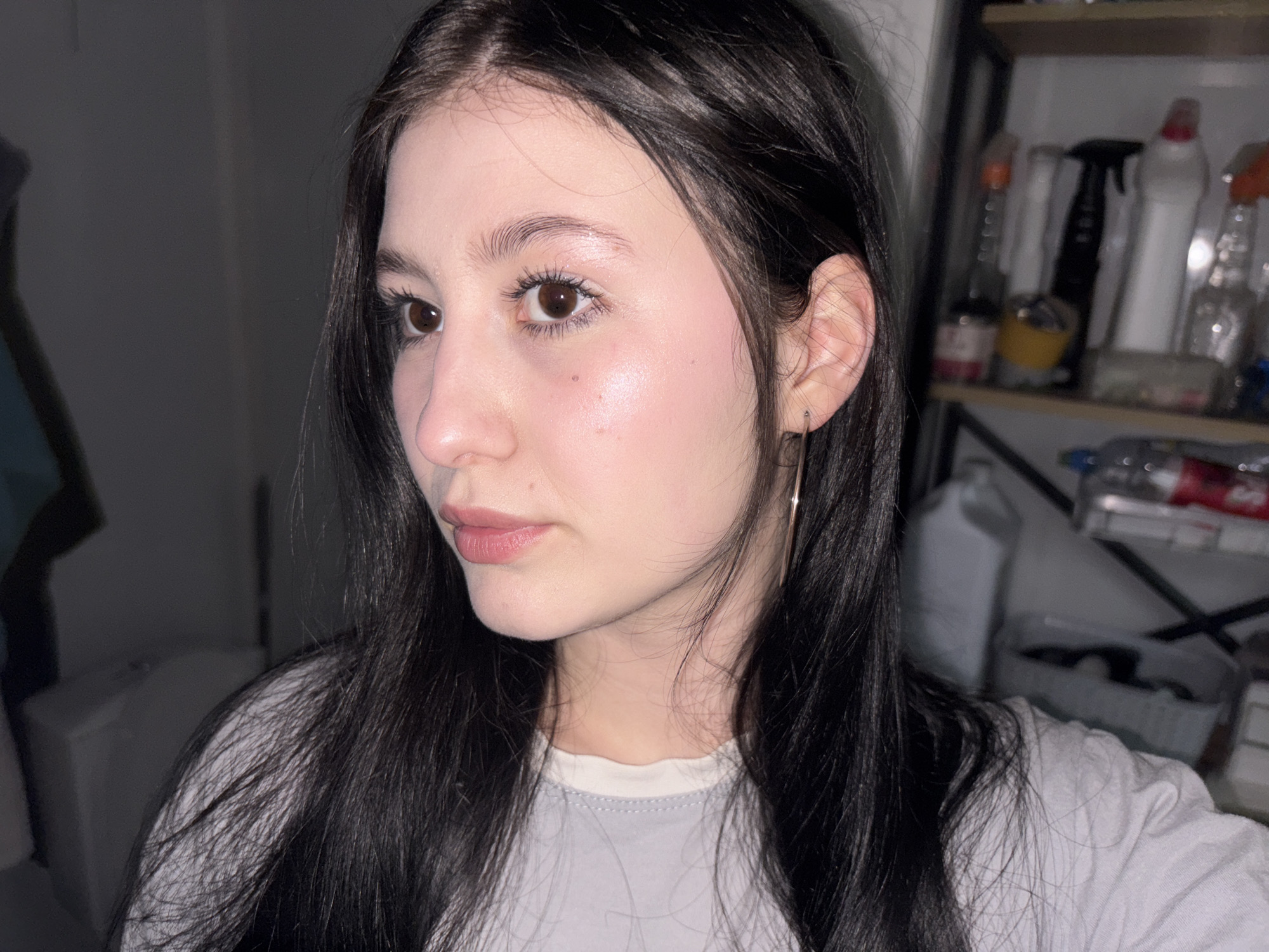 A young woman with dark hair is captured in a close-up, three-quarter profile shot. She has fair skin, brown eyes, and full lips. She is wearing a simple earring. She is wearing a white t-shirt. The background is blurred, suggesting an indoor setting with a shelf containing various household items, including cleaning supplies. The image does not provide any location information about Фоки, Russia.