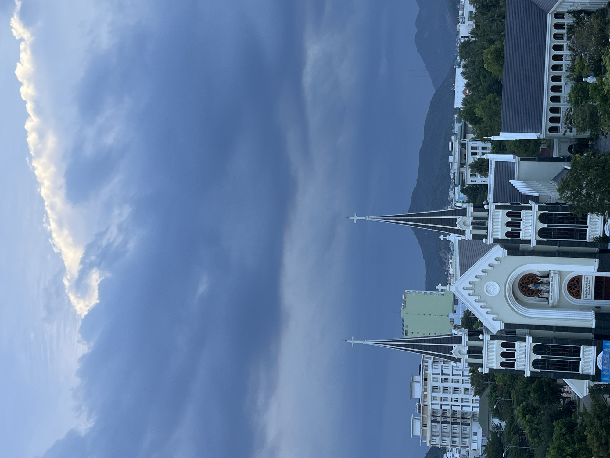 The image depicts a low-angle view in Bac Nha Trang Ward, Vietnam. Two church spires rise towards a predominantly cloudy sky. To the right, buildings and mountains are visible behind the church. The sky shows variations in cloud cover, ranging from light to dark shades.