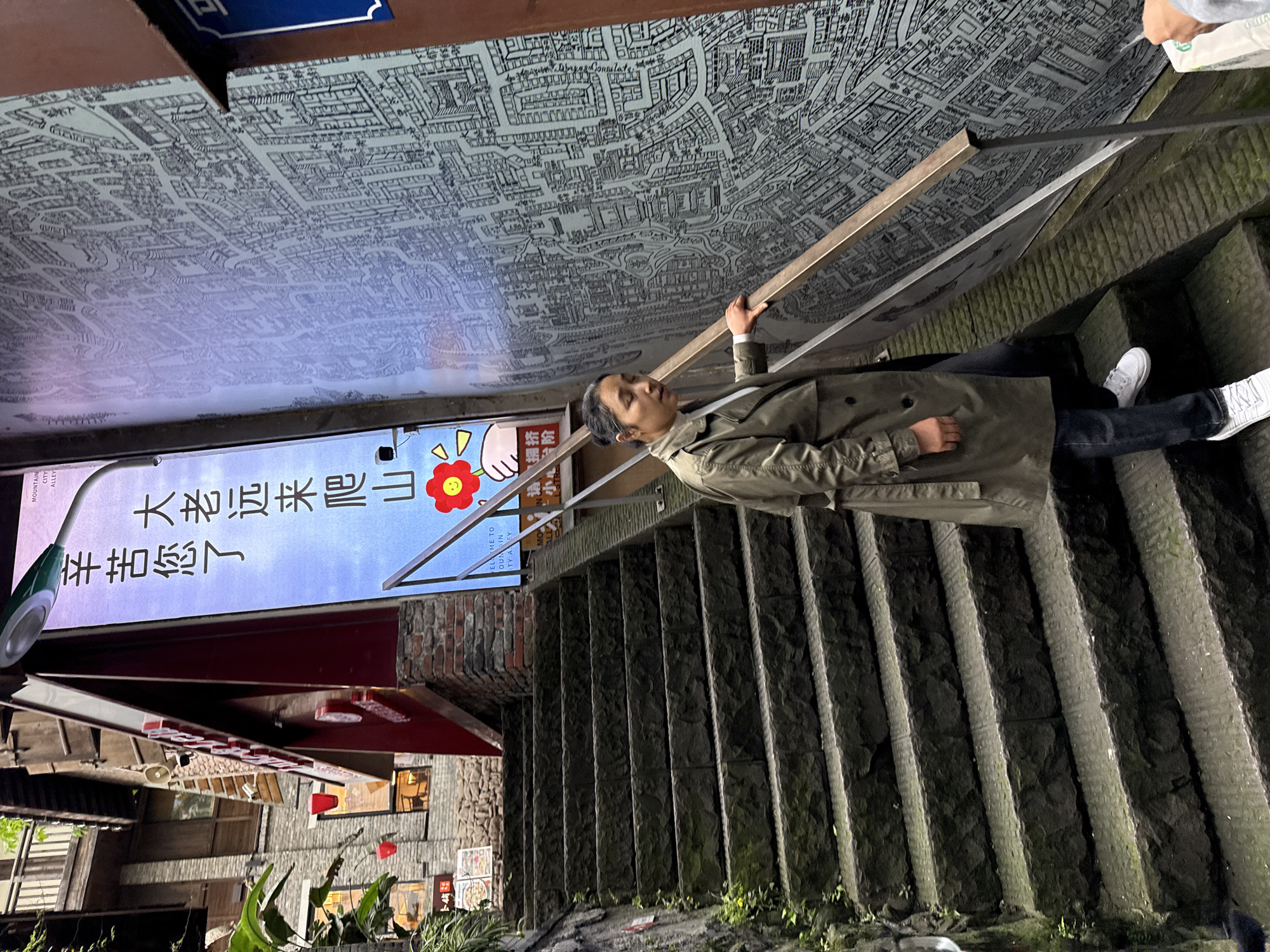 A medium-angle, full-body shot depicts an adult female descending a stone staircase in a narrow urban alleyway within Yuzhong District, China, specifically identified by a sign as "Mountain City Alley." The woman, positioned centrally, wears an olive-green trench coat, a white long-sleeved shirt, dark blue jeans, and white sneakers. Her dark hair is pulled back. She is looking towards the right side of the frame with a slightly open mouth, her right hand resting on a metal handrail with a light-colored top rail.
The staircase is composed of dark, roughly hewn stone blocks, with metal tread inserts on each step. Green moss and small plants adhere to the stone retaining wall on the left. The right wall features a large, black-and-white mural depicting a detailed urban map.
To the left, a tall, vertical illuminated sign displays Chinese characters, "MOUNTAIN CITY ALLEY" in English, and a graphic of a red smiling flower. The Chinese text translates to "You've worked hard, coming so far to climb the mountain." Above this sign, a green and grey streetlamp extends. Further left and in the background, multi-story buildings with traditional wooden balconies, brick facades, and sloped roofs are visible. One building exhibits a prominent red business sign with white Chinese characters and red hanging lanterns.
In the bottom right corner, the right hand and forearm of a second, unseen individual are visible, holding a white bag with a partially obscured green circular logo.