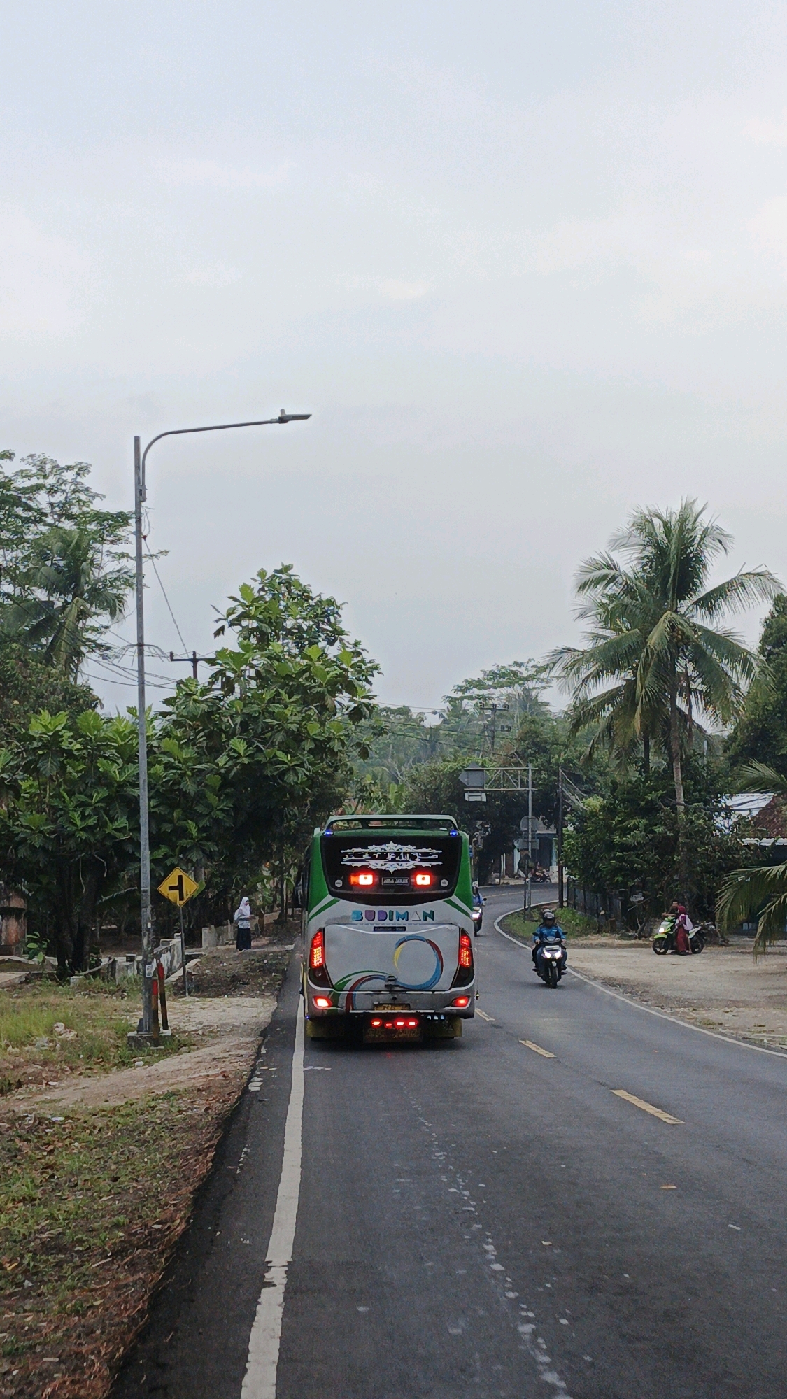 The image captures a view from a vehicle traveling on a paved road in Sukaresik, Indonesia. The sky is overcast and a street lamp stands on the left side of the road. A green and white bus with "BUDIMAN" written on the back is in the foreground, moving away from the viewer. To the right of the bus, a motorcycle with a rider is also moving away. Further down the road, another motorcycle with two people is parked or stopped on the side. The roadside is lined with lush green trees and vegetation. A yellow directional sign is visible on the left, near a person standing by the side of the road. The road has yellow and white lane markings.