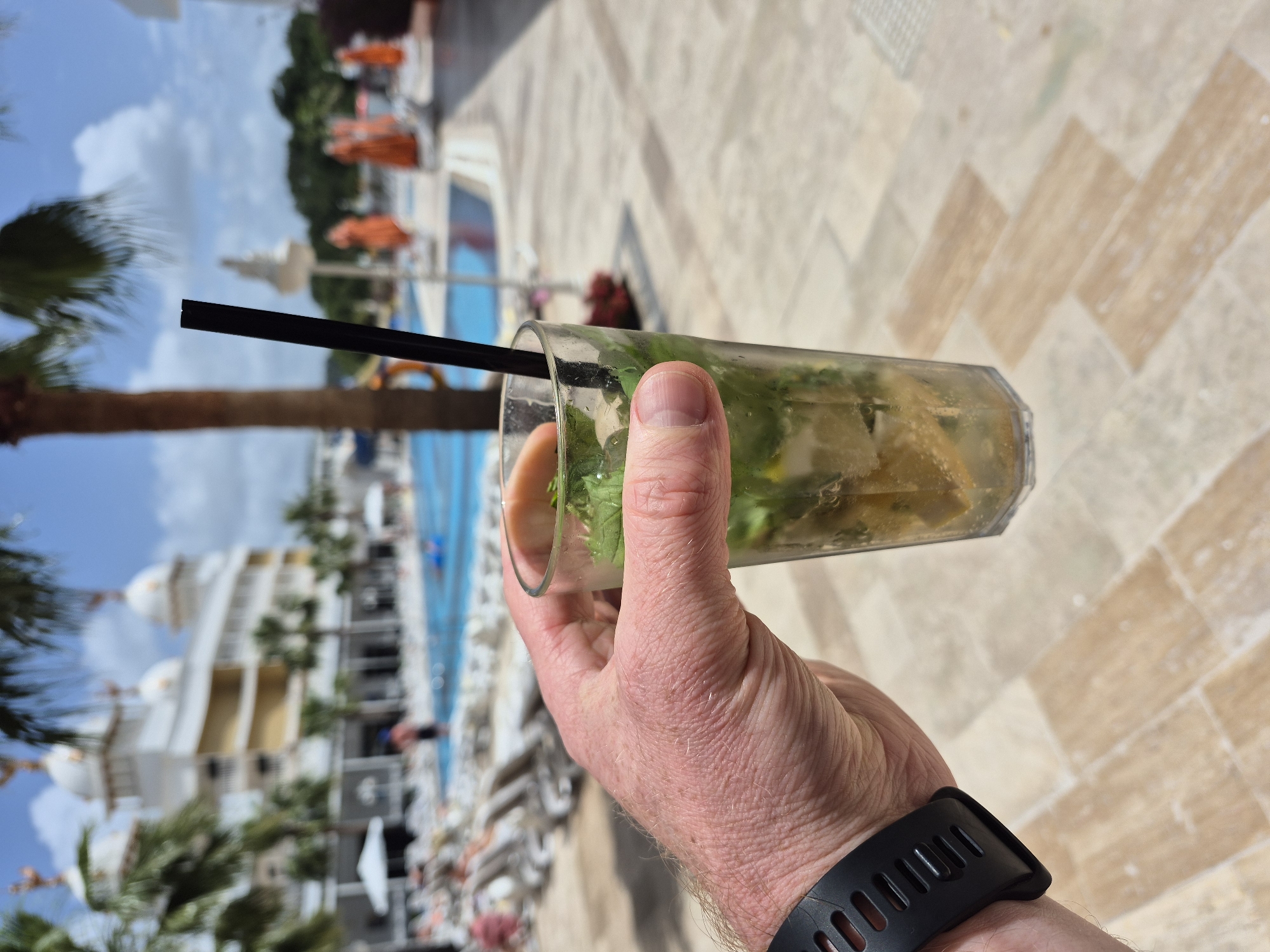 A person holds a tall glass of a mojito-like beverage with a black straw in their right hand. The drink appears to contain ice, mint, and citrus fruit. The person is wearing a black sports watch. The location is a resort in Alanya, Turkey, as evidenced by the architectural style of the white buildings with domes and the presence of palm trees and a swimming pool in the background. Several lounge chairs and people are visible near the pool. The ground is paved with light-colored, geometric tiles. The image is taken in daylight.
