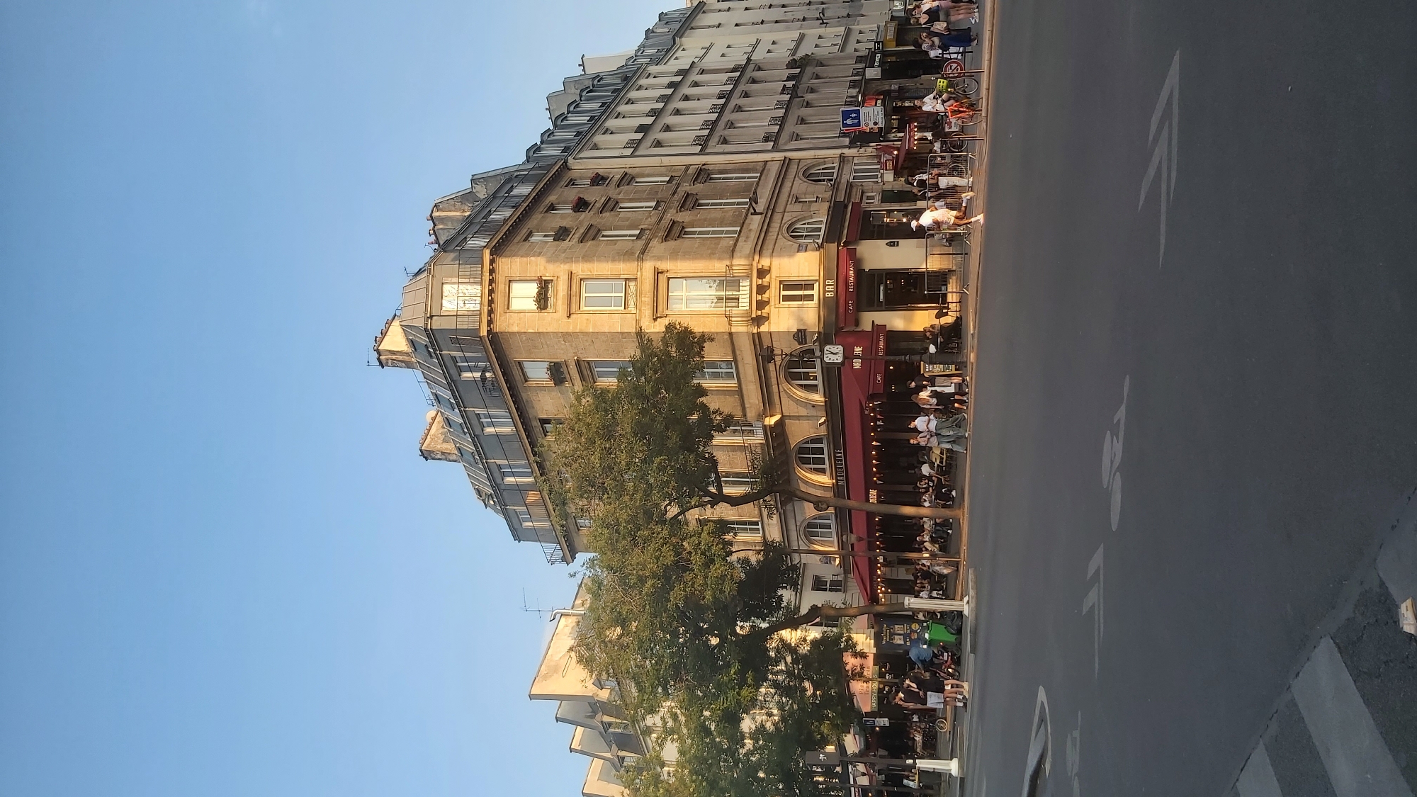The image shows a street scene in Paris, France, on a clear, sunny day. The focal point is a large, corner building with a mansard roof, characteristic of Parisian architecture. The building is made of light-colored stone and has multiple stories with windows and balconies.
At street level, a cafe with a red awning and outdoor seating is busy with patrons. Signs on the building indicate "MADELEINE" and "BAR CAFE RESTAURANT." A large tree with lush green foliage stands in front of the building, providing shade.
Across the street, there are more buildings, some appearing older than the main corner structure. The street is paved with asphalt and features painted markings for a bicycle lane. Several people are visible walking, cycling, and sitting at cafe tables. There are also some electric scooters parked along the sidewalk. The overall atmosphere suggests a lively, urban setting in the late afternoon or early evening, with the warm sunlight casting long shadows.