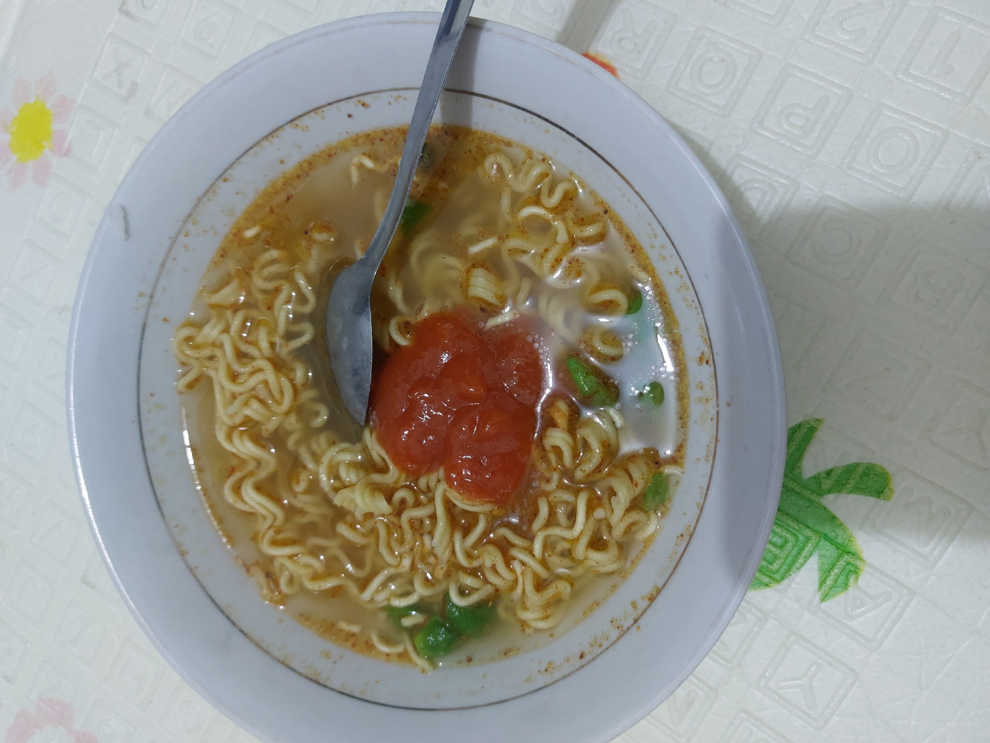 A close-up, overhead shot shows a white bowl of instant noodles on a textured placemat. The noodles are curly and submerged in a light brown broth. A dollop of red chili sauce is placed in the center of the noodles. A silver spoon rests on top of the noodles, partially submerged in the broth. Green pieces of vegetables are also visible in the broth. The placemat has a white textured pattern with faint floral designs in pink and yellow. The scene appears to be indoors, on a table. The specific location in the City of Pematang Siantar, Indonesia, cannot be determined from the image alone.