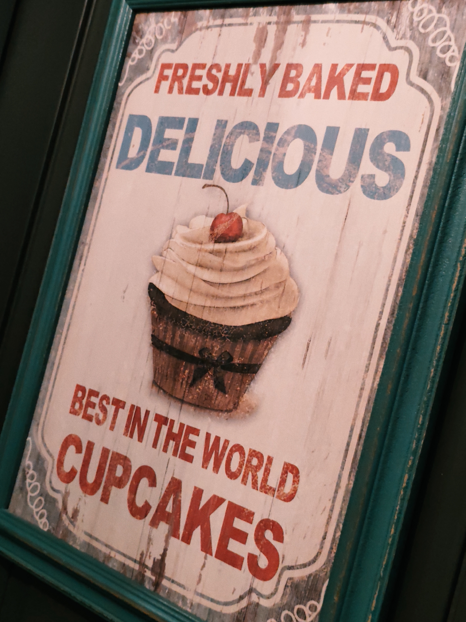 This is a close-up, angled shot of a decorative sign featuring a cupcake illustration. The sign has a vintage, distressed appearance with a wooden texture visible underneath the graphics. A teal-colored frame surrounds the sign.
The main subject is a stylized cupcake with a swirl of cream and a red cherry on top. It sits in a brown wrapper decorated with a black ribbon and bow. The background of the sign is a light, weathered wood pattern.
Visible text on the sign reads: "FRESHLY BAKED", "DELICIOUS", and "BEST IN THE WORLD CUPCAKES". The lettering for "FRESHLY BAKED" is in red, while "DELICIOUS" is in blue and distressed. "BEST IN THE WORLD CUPCAKES" is in red, with "CUPCAKES" being significantly larger.
The lighting appears to be artificial or overcast, creating a soft, diffused light that highlights the texture of the sign and its frame. The angle suggests the sign is hanging or mounted, likely indoors or in a sheltered outdoor location. There are no people visible in the image. The overall impression is that of a charming, retro-style advertisement for cupcakes. The location context is Estosadok, Russia.