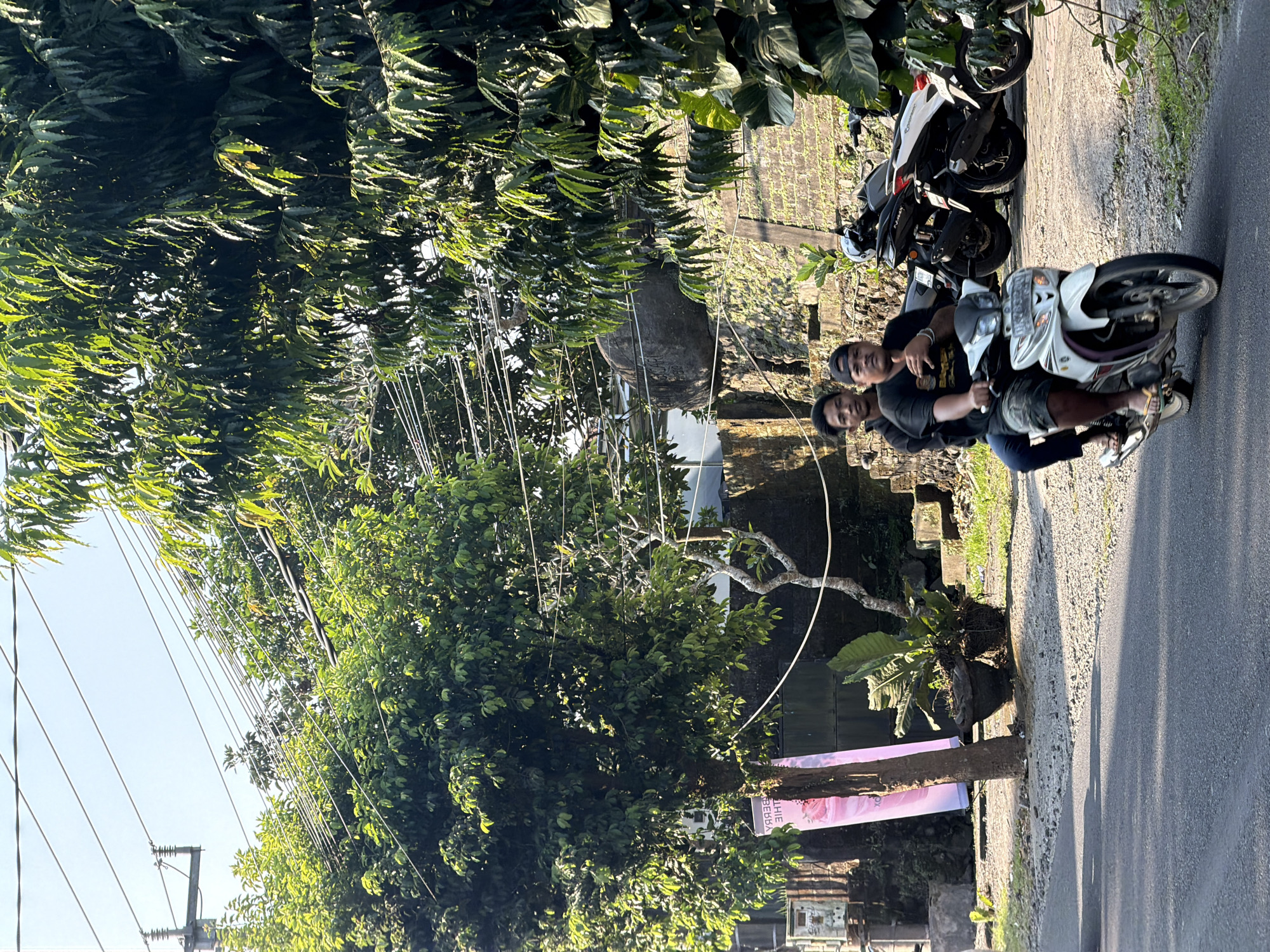 In Ubud District, Indonesia, a white motorcycle carrying two adult males is captured in motion on an asphalt road. The driver, wearing a black t-shirt and patterned shorts, gestures with his left hand while operating the vehicle. The passenger, in a black t-shirt and blue pants, sits behind him. Parked motorcycles, a black and a white scooter, are visible on the right side of the road. The background features lush green foliage, trees with dense canopies, power lines, and stone structures. A pink banner, partially obscured by a tree, stands on the left side of the road. The scene is illuminated by bright sunlight, casting shadows on the road.