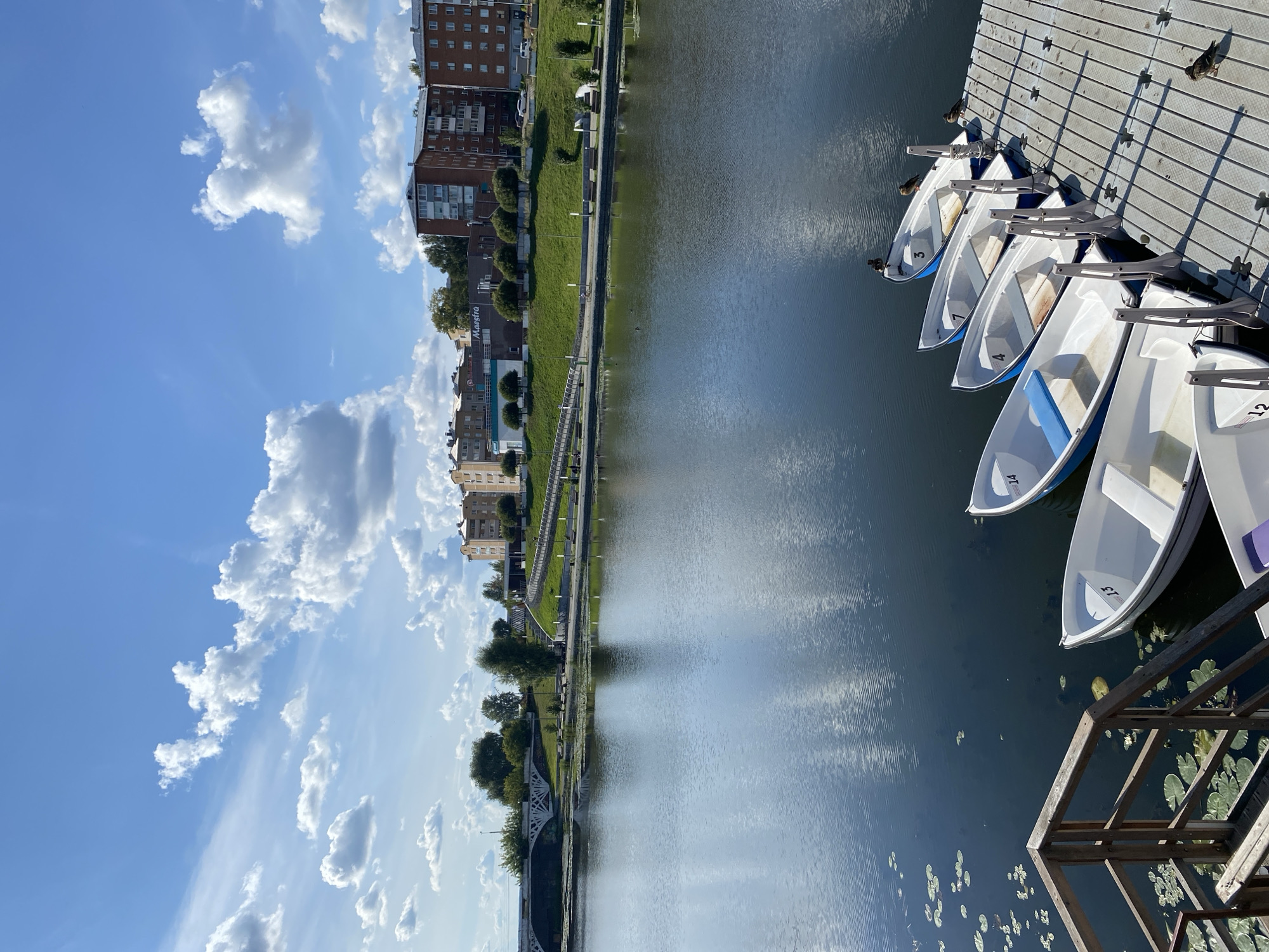 The image depicts a waterfront scene in the city of Votkinsk, Russia. In the foreground, several white rowboats with blue seats are docked at a wooden pier. Some of the boats are numbered, with visible numbers including 14, 13, 7, 4, and 3. Ducks are perched on some of the boats. The water is dark and calm, with lily pads floating on the surface near a wooden railing on the left side of the frame.
In the background, a promenade with benches and lampposts runs along a grassy slope. Beyond the promenade, a modern white bridge with a decorative arch spans the water. Further in the distance, there are multi-story residential buildings and trees. The sky is bright blue with scattered white clouds, and the sun is casting shadows from the pier and the boats onto the water.