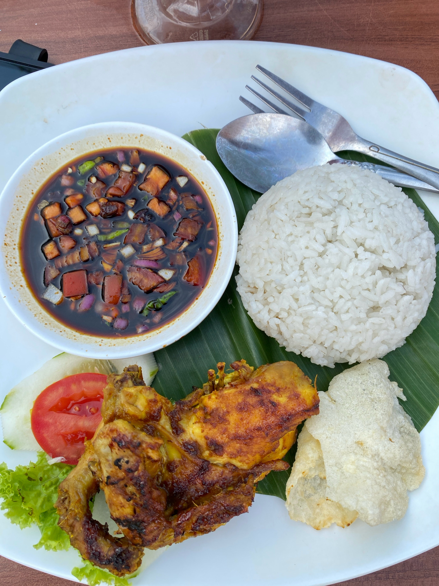 The image shows a close-up, overhead view of a plate of Indonesian food, likely taken at a restaurant in Lancok, Indonesia. The plate, which is white, sits on a wooden surface. In the upper left of the plate is a white bowl filled with a dark sauce containing chopped vegetables and meat. To the right of the sauce is a mound of white rice, placed on a green banana leaf. Below the rice and sauce, a piece of grilled chicken is positioned on the plate, accompanied by a slice of tomato, a lettuce leaf, and a cucumber slice on the left side. On the right side, next to the chicken, are two pieces of light-colored, crispy crackers. A spoon and fork are resting on the plate, above the rice. The lighting suggests it is daytime, and the overall presentation indicates a meal setting.
