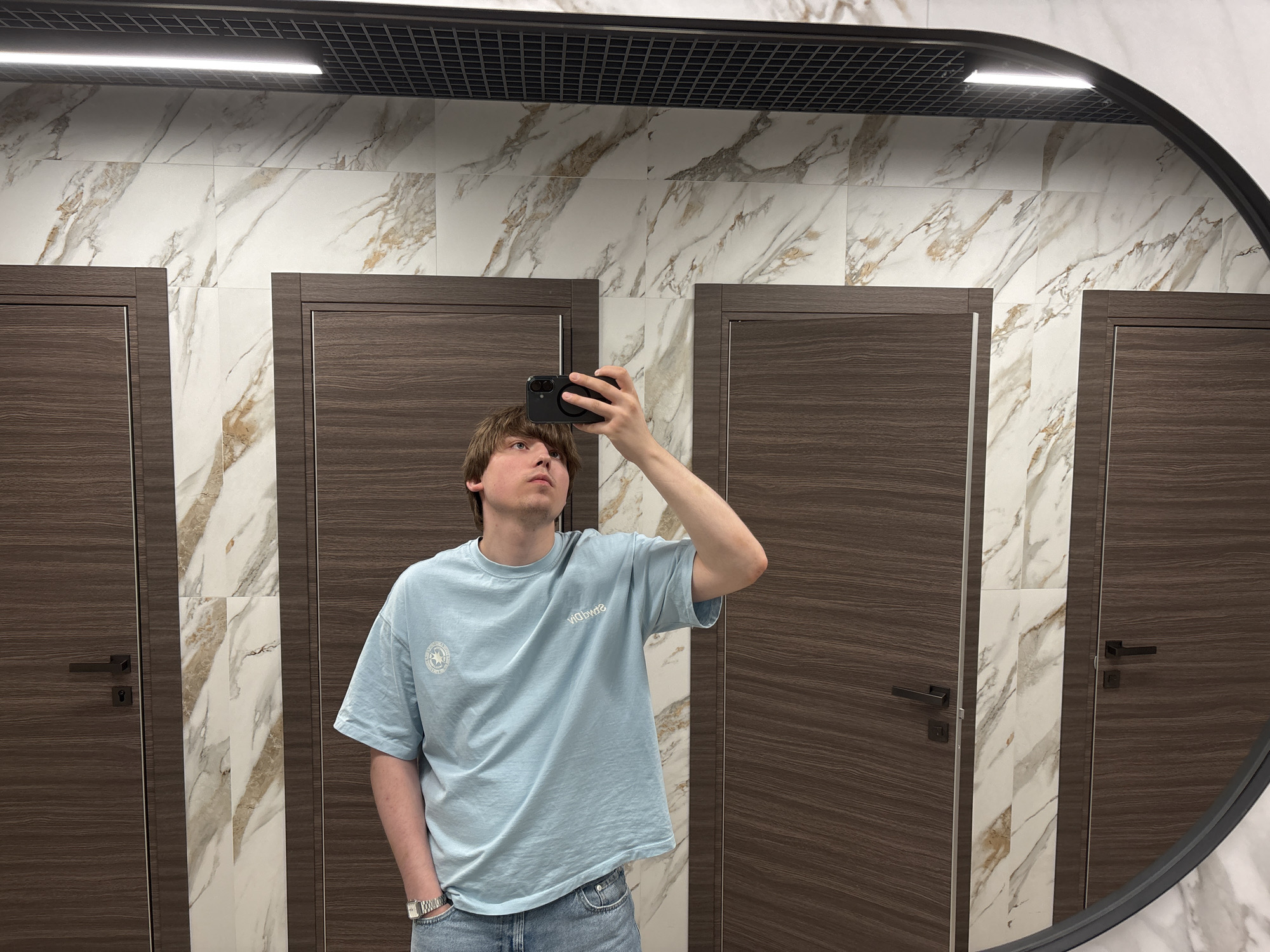 A young man with light brown hair, wearing a light blue t-shirt and blue jeans, is taking a selfie in a restroom with marble-patterned walls and dark wooden doors. He is standing in front of three doors, with the middle door partially obscured by a vertical marble tile accent. The man is holding a black smartphone in his right hand, positioned to capture his reflection in a large, oval mirror. His left hand is in his jeans pocket, and he is looking up towards the mirror. A silver wristwatch is visible on his left wrist. The restroom appears to be modern and well-lit, with a grid-like ceiling featuring a strip light. This scene is located in the city of Dnipro, Ukraine.