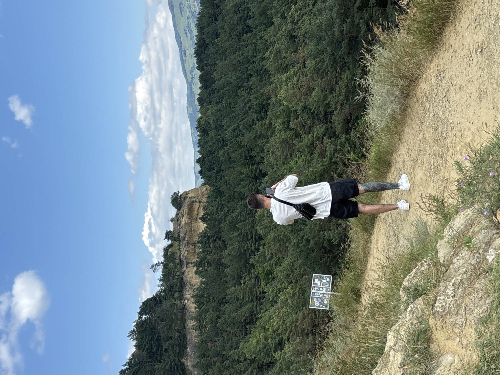 A man stands on a dirt path, facing away from the camera, holding a smartphone to his face as if taking a photo or video. He is wearing a white t-shirt, black shorts, white sneakers, and a black fanny pack slung across his chest. The path is bordered by dry grass and rocks. In the background, a dense forest of pine trees covers rolling hills, and a large, rocky cliff face is visible. The sky is blue with scattered white clouds. To the left of the man, on the path, is a white signpost with multiple panels displaying various symbols and text. There is no indication that this scene is located in Mirny, Russia.