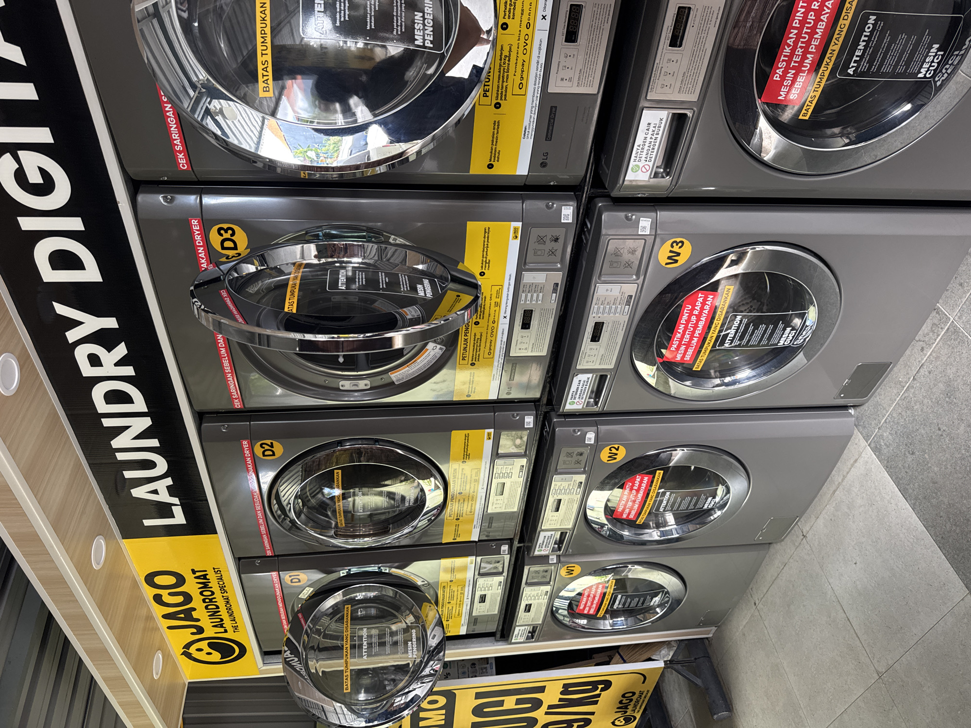 The image shows a stack of commercial laundry machines, likely located in a laundromat in an unidentified city. The machines are gray and labeled with identifiers like "D1," "D2," "D3," "W1," "W2," and "W3." The machines on the left are dryers ("MESIN PENGERING"), while those on the right are washing machines ("MESIN CUCI"). There are various warning and instruction labels in Indonesian on each machine, mentioning detergent usage, door closure, and suggested load limits. A sign bearing the logo of "JAGO LAUNDROMAT" and "LAUNDRY DIGITAL" is visible, along with other signs advertising payment options. The floor appears to be tiled.