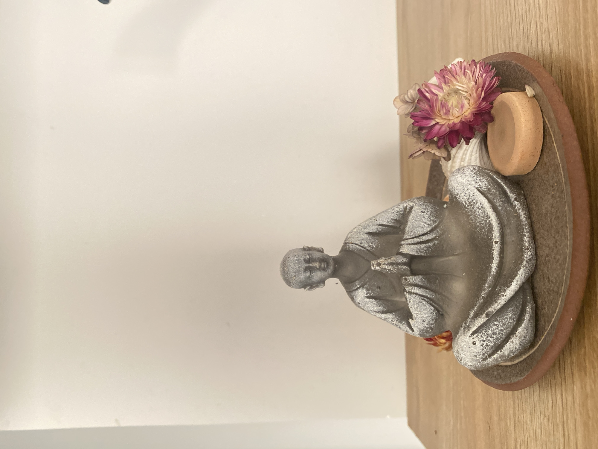 A statue of a person in a meditative pose, possibly a monk or spiritual figure, is placed on a wooden surface near a white wall. The statue has a textured, possibly stone-like appearance, and is positioned in a decorative brown tray. A pink and white flower is placed behind the statue, and a small, round, light-brown object sits on the right side of the statue in the tray. The setting appears to be indoors, perhaps in a home or meditative space. The location of Lâm Viên - Đà Lạt, Vietnam, cannot be verified from the image.