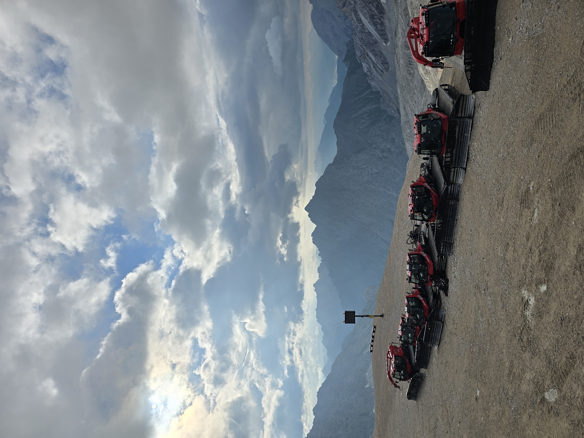 The image depicts a scenic mountain landscape with several red snowcats lined up on a gravelly plateau. In the background, a vast expanse of layered mountain ranges stretches towards the horizon under a dramatic sky filled with clouds. The snowcats, which appear to be parked and not in operation, are the primary subjects in the foreground and midground. A signpost with a digital display is visible behind the row of snowcats, and further back, two benches are placed on the plateau. The overall environment suggests a high-altitude location, possibly a ski resort or a mountain pass, given the presence of snow grooming vehicles and the rugged terrain. The location is likely on the Zugspitze, Germany's highest peak, which is near Garmisch-Partenkirchen.
