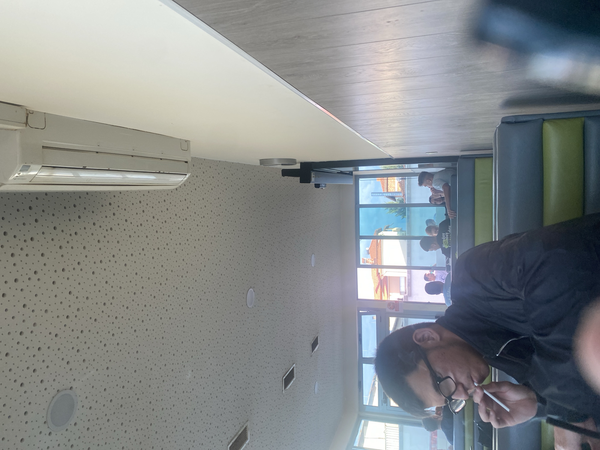 In Ablanitsa, Bulgaria, a young man wearing glasses and a black jacket is seated at a booth, smoking. He is facing to the left and appears to be in a restaurant or similar establishment. The ceiling is white and perforated with numerous small holes, and there is a white air conditioning unit mounted on the wall. The walls are paneled with light-colored wood-like material. Through a large window, several other people are visible, as are buildings with red tile roofs. The seating in the foreground consists of gray and green upholstered booths.