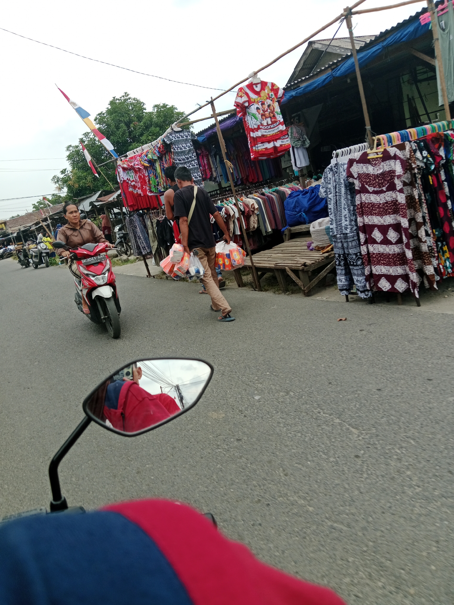 In the city of Binjai, Indonesia, on a cloudy day, an individual on a red scooter is captured in the foreground, their reflection visible in the scooter's side mirror. The reflection shows the rider wearing a red jacket and blue clothing. The scooter's license plate reads "BK 3024 XNL". The scooter is moving along a paved road, with a market stall set up on the right side of the road.
The market stall displays a variety of clothing items, including shirts and dresses, hanging on racks. Several people are present at the market. A woman in a brown jacket is riding another scooter in the background, and a man in a black shirt and beige pants is walking past the market stall, carrying several shopping bags. Two flags, one larger than the other, are visible in the upper left corner of the image, seemingly attached to a pole. The market appears to be located along a busy street, with other vehicles and structures visible in the distance.