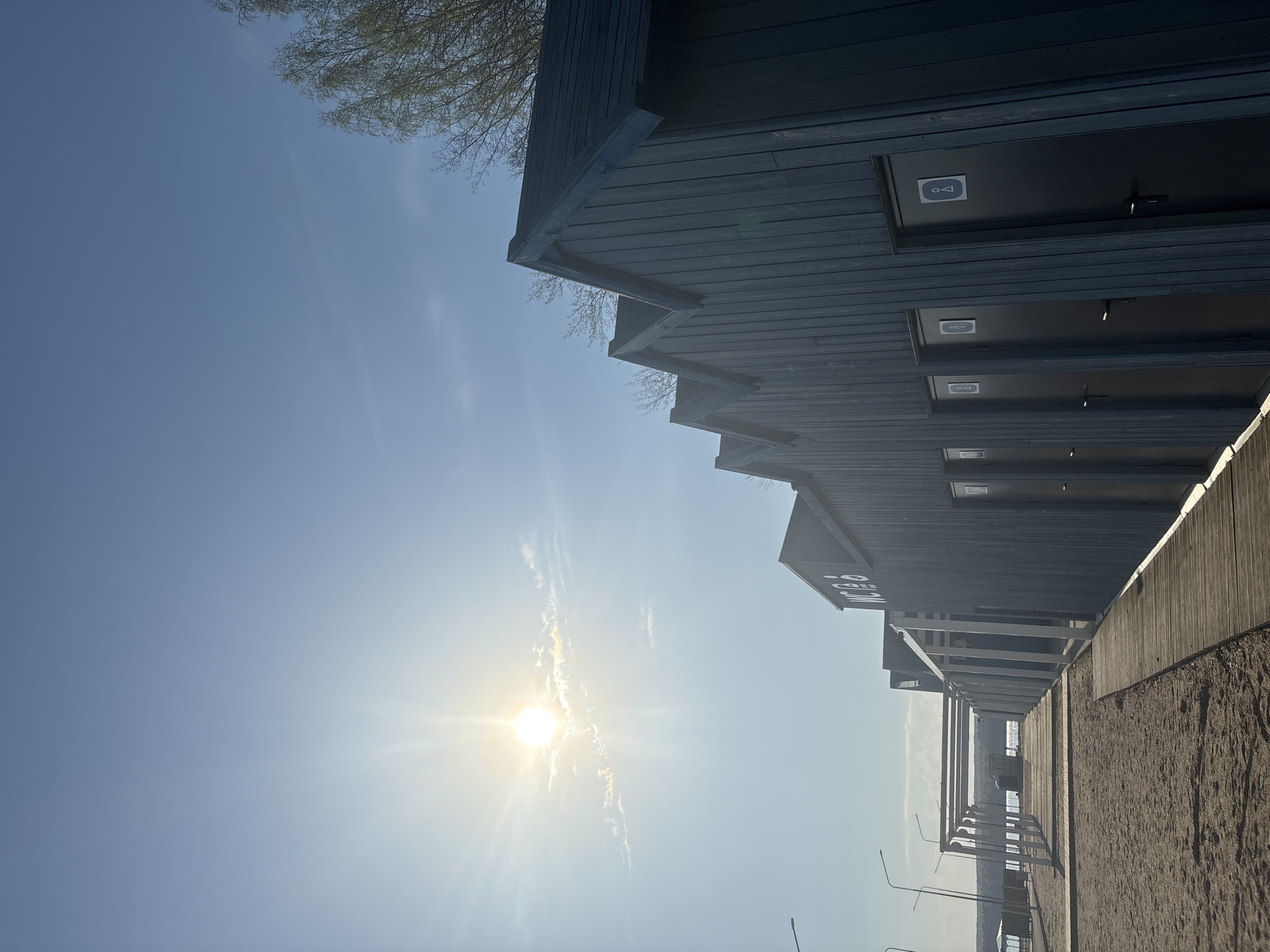 The image depicts a series of modern wooden structures with a distinctive sawtooth roofline, likely public restrooms, as indicated by the restroom symbol on one of the doors. These structures are located in the city of Chaikovsky, Russia, near a sandy area, possibly a beach or riverbank. The sun is bright in the sky, casting shadows and creating a lens flare effect. A line of streetlights is visible further down the path. A glimpse of what might be a body of water and the horizon is present between the structures. Bare branches of a tree are visible in the upper left corner of the image.
