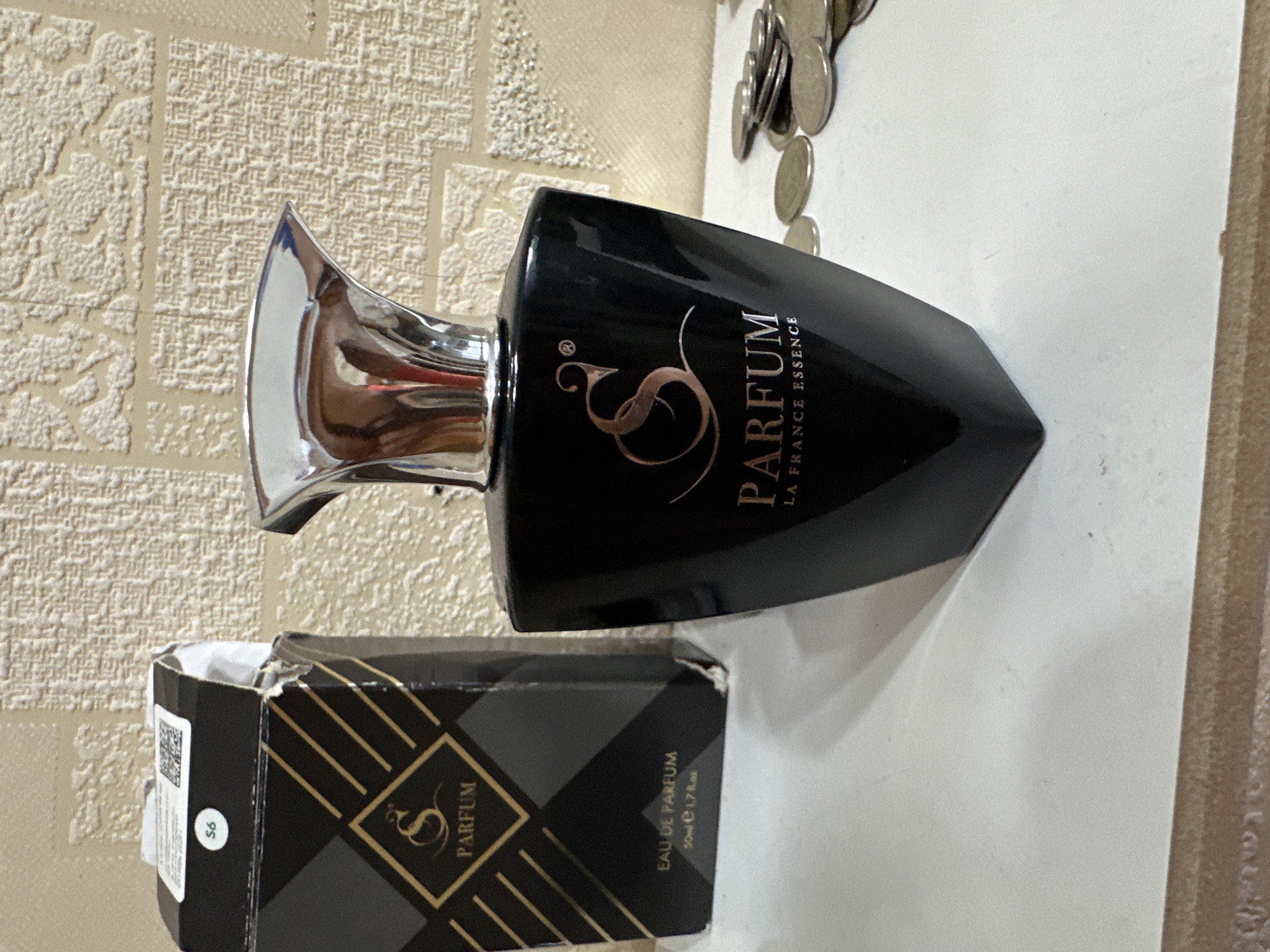 A black bottle of perfume with a silver cap is placed on a white surface. The bottle has a stylized "S" logo and the words "PARFUM LA FRANCE ESSENCE" printed on it in rose gold. Next to the bottle is its partially open black and gold cardboard box, which also displays the "S" logo and "PARFUM". The background is a textured, light-colored wallpaper. A pile of coins is scattered to the right of the perfume bottle. The image appears to be taken indoors, likely in a domestic setting. There is no visible indication of its location in Rostov-on-Don, Russia.