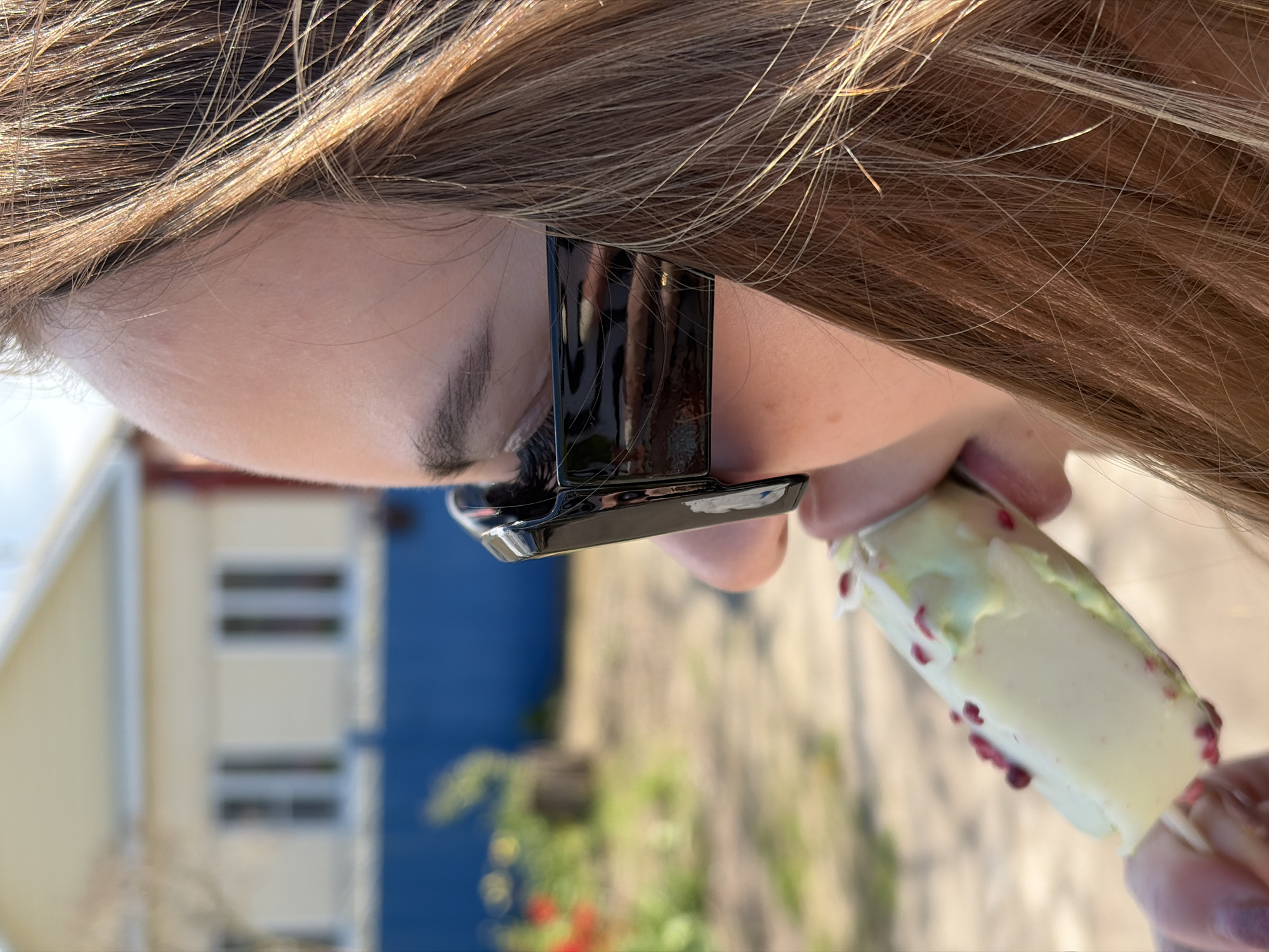 A close-up shot shows a person, presumably female, with light brown hair and fair skin, eating a white-coated popsicle with red specks. They are wearing black rimmed glasses. The background suggests an outdoor setting. A building with a yellow exterior and a blue wall is visible. The surroundings appear to include vegetation, possibly in a garden or yard. Without additional information, it is impossible to confirm if this image was taken in Port-Katon, Russia.
