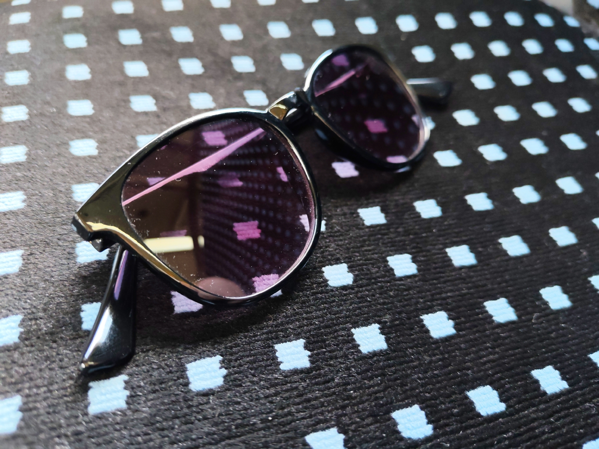 The image shows a pair of sunglasses resting on a textured surface. The sunglasses have black frames with a gold accent on the bridge and purple tinted lenses. The surface is black with a pattern of light blue squares. The location and city are unknown.