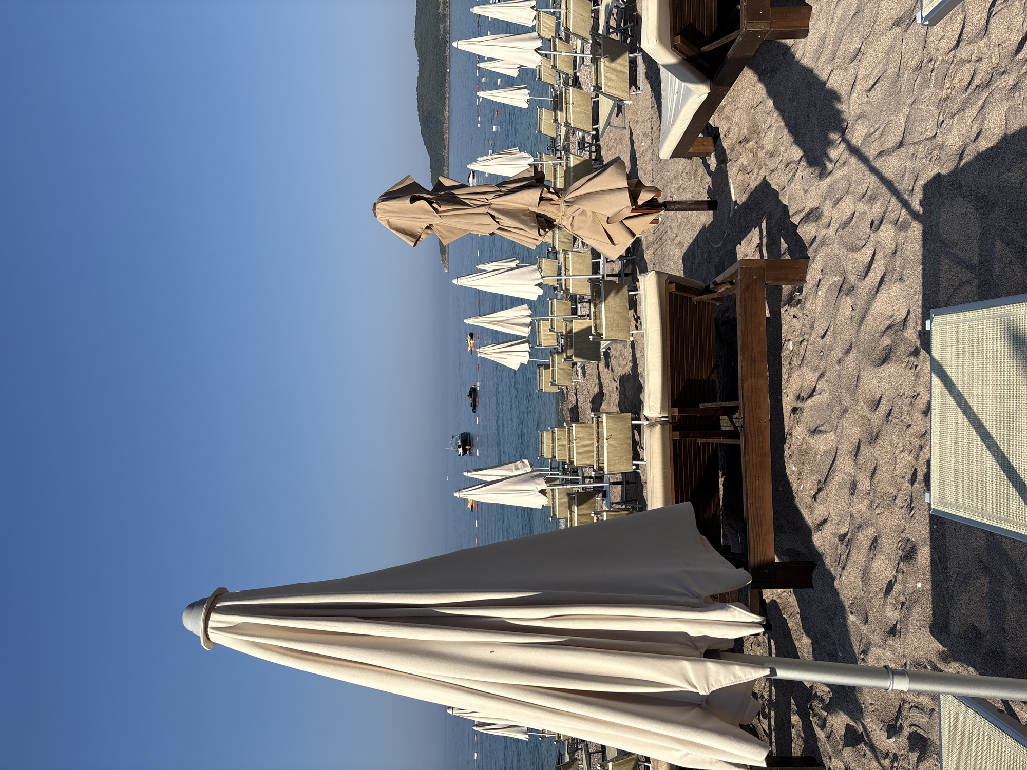 The image depicts a serene beach scene in Kamenovo, Montenegro, on a clear, sunny day. The foreground is dominated by closed beach umbrellas and rows of empty lounge chairs neatly arranged on the sandy shore. The umbrellas are of two types: some are tall, closed, and pale beige, while another in the middle is a taupe color, folded and tied. The lounge chairs are made of wood and fabric. Beyond the beach, the calm blue sea stretches towards the horizon, with a few boats and jet skis visible. The background features a gentle slope of green hills covered in vegetation. The overall atmosphere is one of quiet tranquility, suggesting the early morning or late afternoon when the beach is not yet crowded.