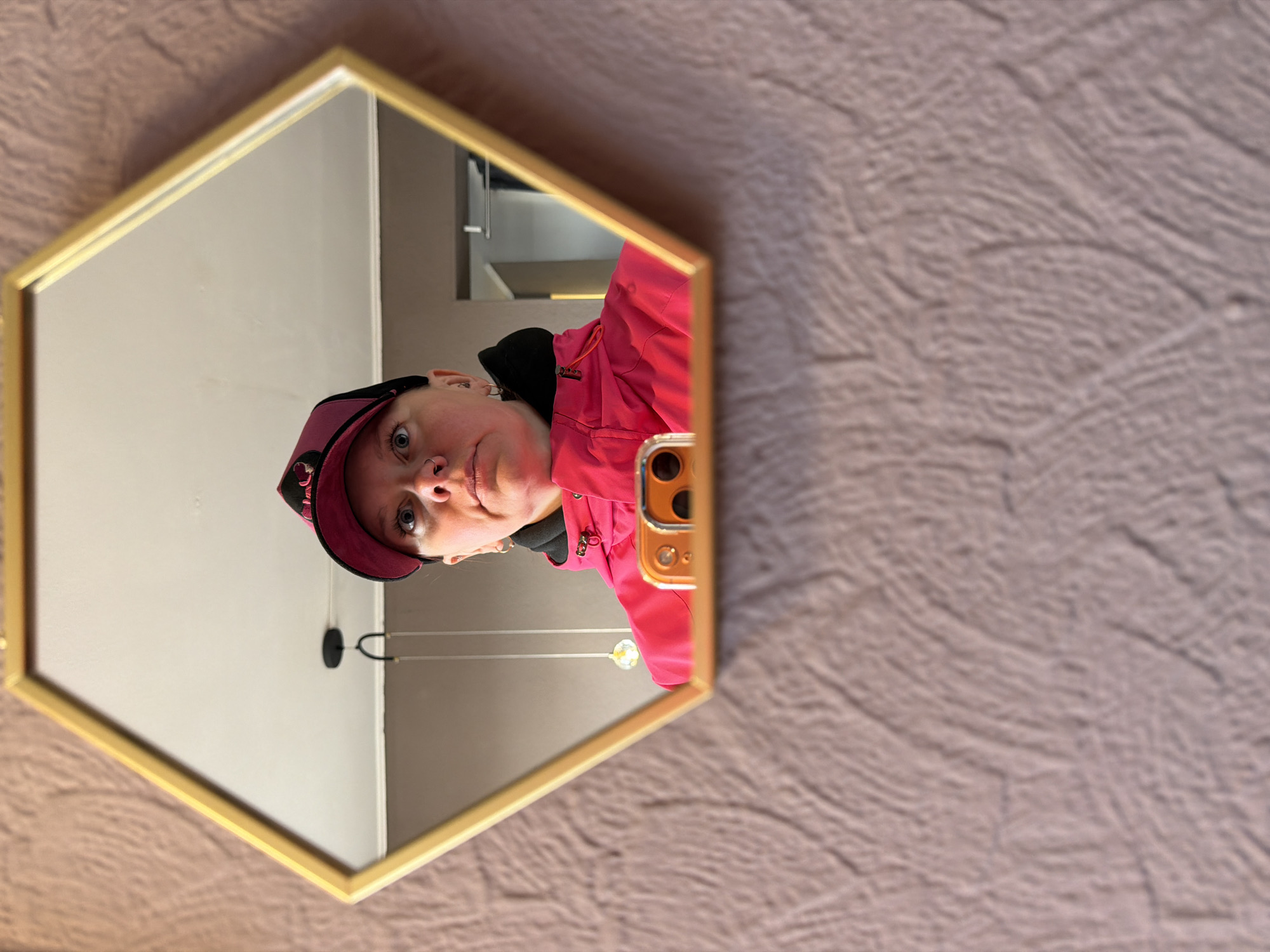 The image captures a mirror selfie, presented rotated 90 degrees counter-clockwise from its likely original vertical orientation. A person, appearing female, is reflected in a hexagonal mirror with a gold-colored frame. She is wearing a bright pink jacket and a dark cap with pink detailing. Her expression is neutral, and her blue eyes are wide open, looking directly towards the camera, with a visible nose piercing.
The person is holding a smartphone, identifiable by its gold or orange casing and visible multi-lens camera module, positioned to capture the reflection. Inside the mirror, behind the person, a plain, light-colored wall is visible, along with a modern pendant light fixture featuring two suspended bulbs. The wall surrounding the mirror, outside its frame, is textured with an undulating, wave-like pattern, rendered in a soft brownish-pink or taupe color, suggestive of wallpaper or a decorative paint finish. The scene takes place indoors in Chaikovsky, Russia. There is no visible text in the image.