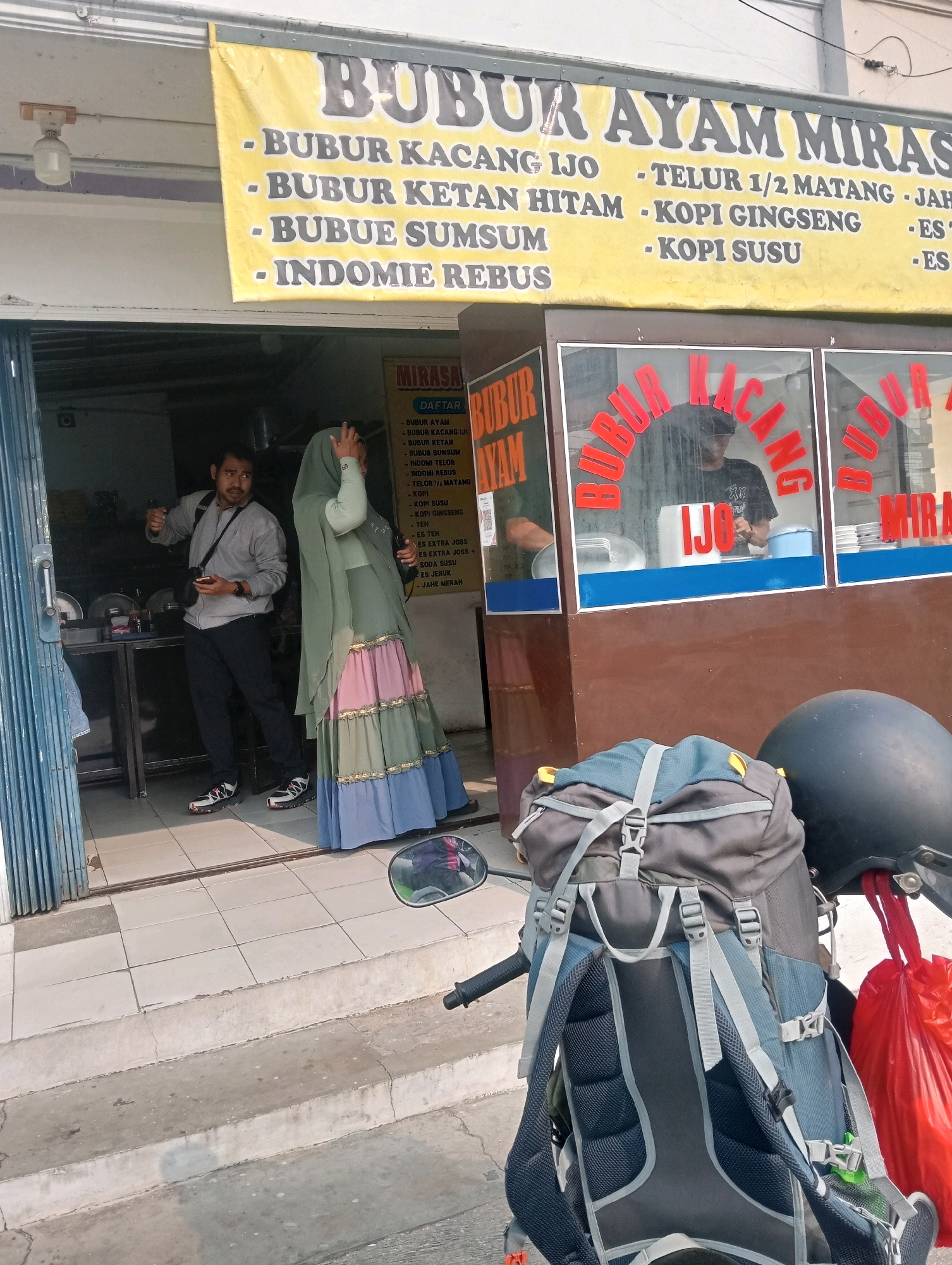 This is a medium shot taken outdoors in what appears to be Bandar Lampung, Indonesia, showing the exterior of a small food stall. A yellow banner with red and black text hangs above the entrance, advertising "BUBUR AYAM MIRASA" (Mirasa Chicken Porridge) and listing various menu items like "BUBUR KACANG IJO" (Mung Bean Porridge), "BUBUR KETAN HITAM" (Black Sticky Rice Porridge), "BUBUR SUMSUM," "INDOMIE REBUS" (Boiled Indomie Noodles), and beverages such as "KOPI GINGSENG" (Ginseng Coffee) and "KOPI SUSU" (Milk Coffee).
In the doorway, a man in a grey jacket and black pants is looking to his left, gesturing with his right hand. He is holding a smartphone in his left hand. Beside him stands a woman wearing a light green hijab and a long, layered dress in shades of green, pink, and blue. She has her right hand raised to her head.
Through the glass window of the stall, a person in a dark t-shirt and cap is visible, appearing to be preparing food. Red lettering on the window also spells out "BUBUR KACANG IJO."
In the foreground, a large backpack, primarily grey and blue, is mounted on the back of a motorcycle. A red plastic bag is hanging from the motorcycle's handlebars. The ground is tiled, with concrete steps leading up to the entrance. The lighting suggests it is daytime.