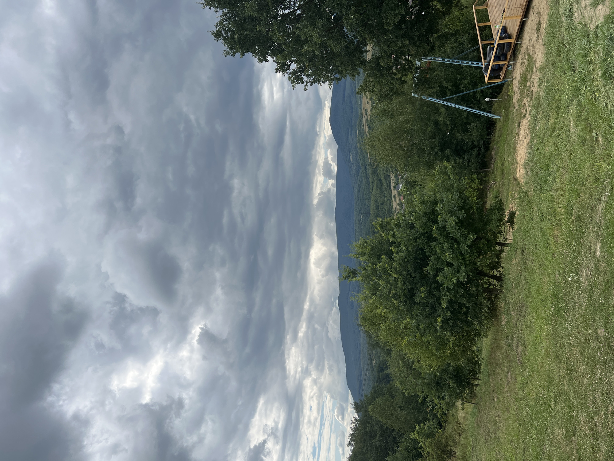 This is a landscape photograph taken in a rural area, possibly near the city of Сонячне, Ukraine. The image is dominated by a dramatic, cloudy sky with heavy, dark gray clouds overhead. Below the sky, a range of rolling, tree-covered hills recedes into the distance, with a lighter, hazy mountain range visible further back. In the foreground, a grassy slope leads down to a wooded area with lush green trees. To the right of the image, a wooden platform is visible, with a metal structure that appears to be part of an outdoor attraction, possibly a zipline or swing set, with several black tires stacked on the platform. The overall impression is one of a natural, outdoor setting under an overcast sky, with signs of recreational activity. There are no people or animals visible in the image.