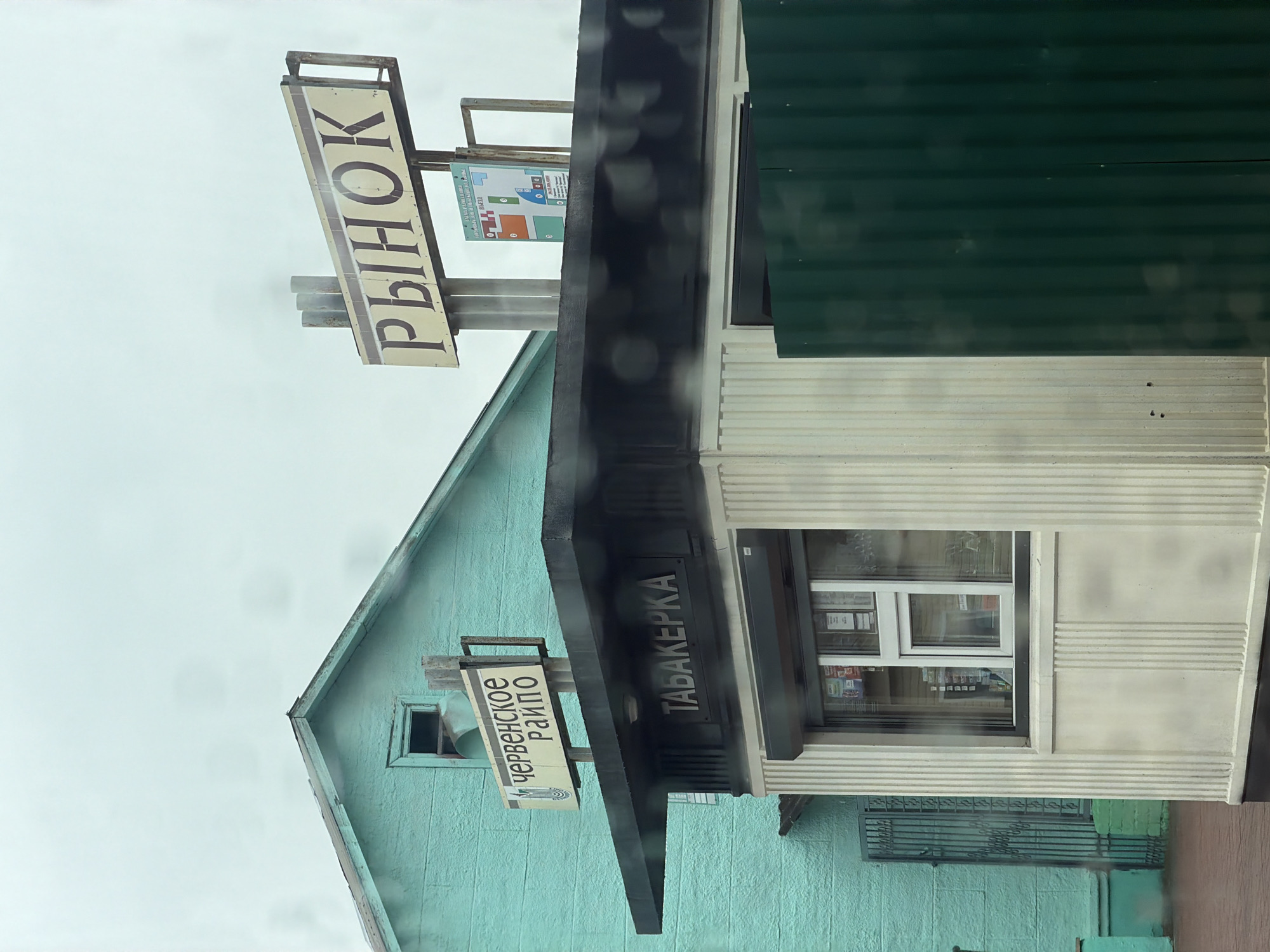 The image shows a small, light-colored kiosk with vertical fluted columns on the front, positioned in front of a teal-colored building. Above the kiosk, a sign reads "ТАБАКЕРКА" in dark letters. To the left of the kiosk, a portion of the teal building is visible, featuring a dark metal gate. Above the teal building, a sign displays "ЧЕРВЕНСКОЕ РАЙПО". Further to the right and higher up, a larger sign reads "РЫНОК" (market) in large, dark letters on a beige background. A map is also attached to the pole supporting the "РЫНОК" sign. The sky appears overcast. The overall scene suggests an outdoor market or commercial area, likely in the city of Cherven, Belarus, given the signage.
