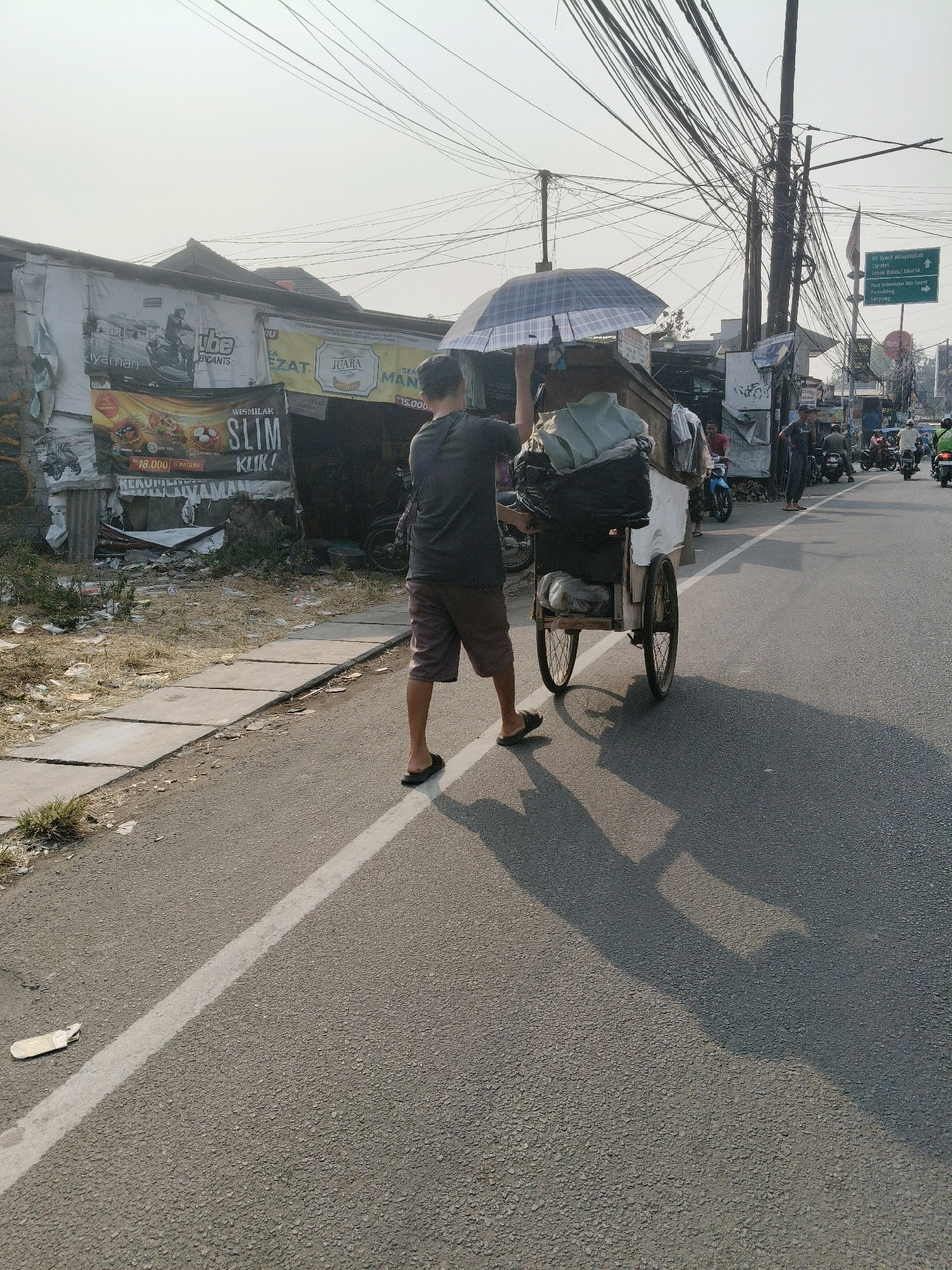The image depicts a man pushing a cart on a street in South Tangerang, Indonesia. The cart is laden with various items, covered by a blue and white checkered umbrella. The man is walking away from the camera, dressed in a dark t-shirt and shorts, with his shadow cast on the asphalt road. To the left of the road, there is a sidewalk with scattered debris and a few buildings adorned with advertisements. The background reveals a complex network of overhead wires and utility poles, characteristic of an urban Indonesian setting. Several motorcycles are visible on the road, suggesting active traffic. A directional signpost indicates "RS Syarif Hidayatullah Ciputat, RS Pondok Indah, BSD/Jakarta, Pondok Petir, Serpong." The scene appears to be set during daylight hours, with a bright, hazy sky.