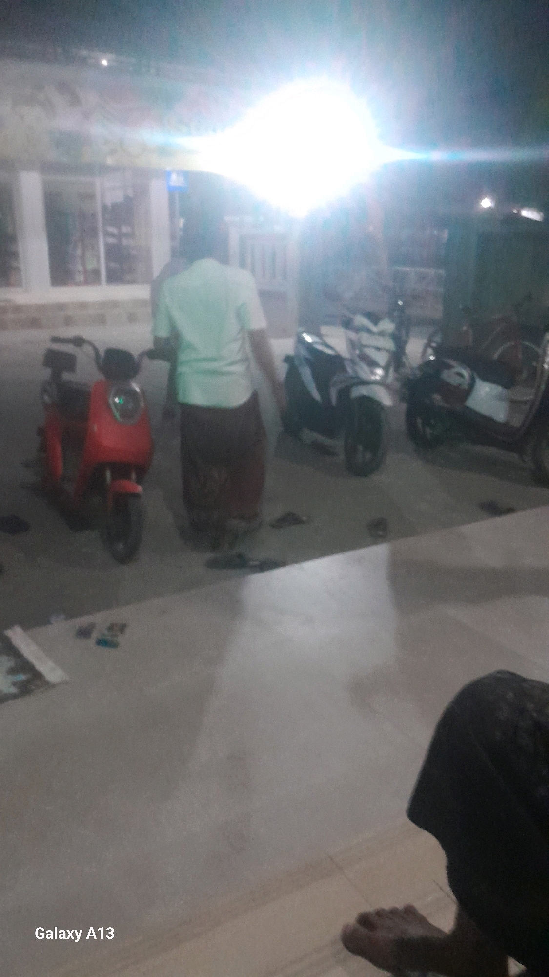 The image depicts an outdoor scene at night, possibly in a public area or near a building in Ngelo, Indonesia. A person is standing between two scooters. One scooter is red and the other appears to be white and black. The person is wearing a light-colored t-shirt and a dark, patterned skirt or sarong. The area is lit by a bright light source, possibly a streetlamp or a flash, which causes overexposure in some parts of the image. The ground is paved, and there are other indistinct objects and shadows visible. In the foreground, a person's foot and leg are visible, indicating that the photo was taken from a seated or low vantage point. The text "Galaxy A13" is visible in the bottom left corner, suggesting the device used to take the picture.
