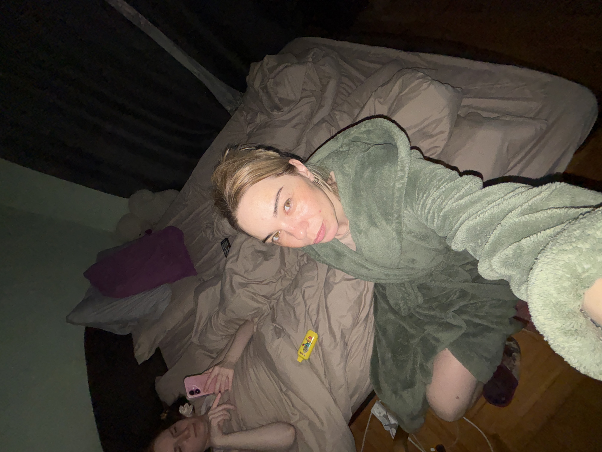 The image shows two individuals in a bedroom. One individual, likely the photographer, is wearing a green robe and holding the camera at an angle, capturing a selfie. Their complexion appears light with a few freckles. The second individual is lying on the bed beneath a beige comforter, holding a pink smartphone. The room features a bed covered with beige bedding and pillows, including a purple decorative pillow. A yellow bottle of some sort is on the bed as well. The room appears to be dimly lit, with dark curtains visible in the background. The location in the city is Unknown, Unknown.