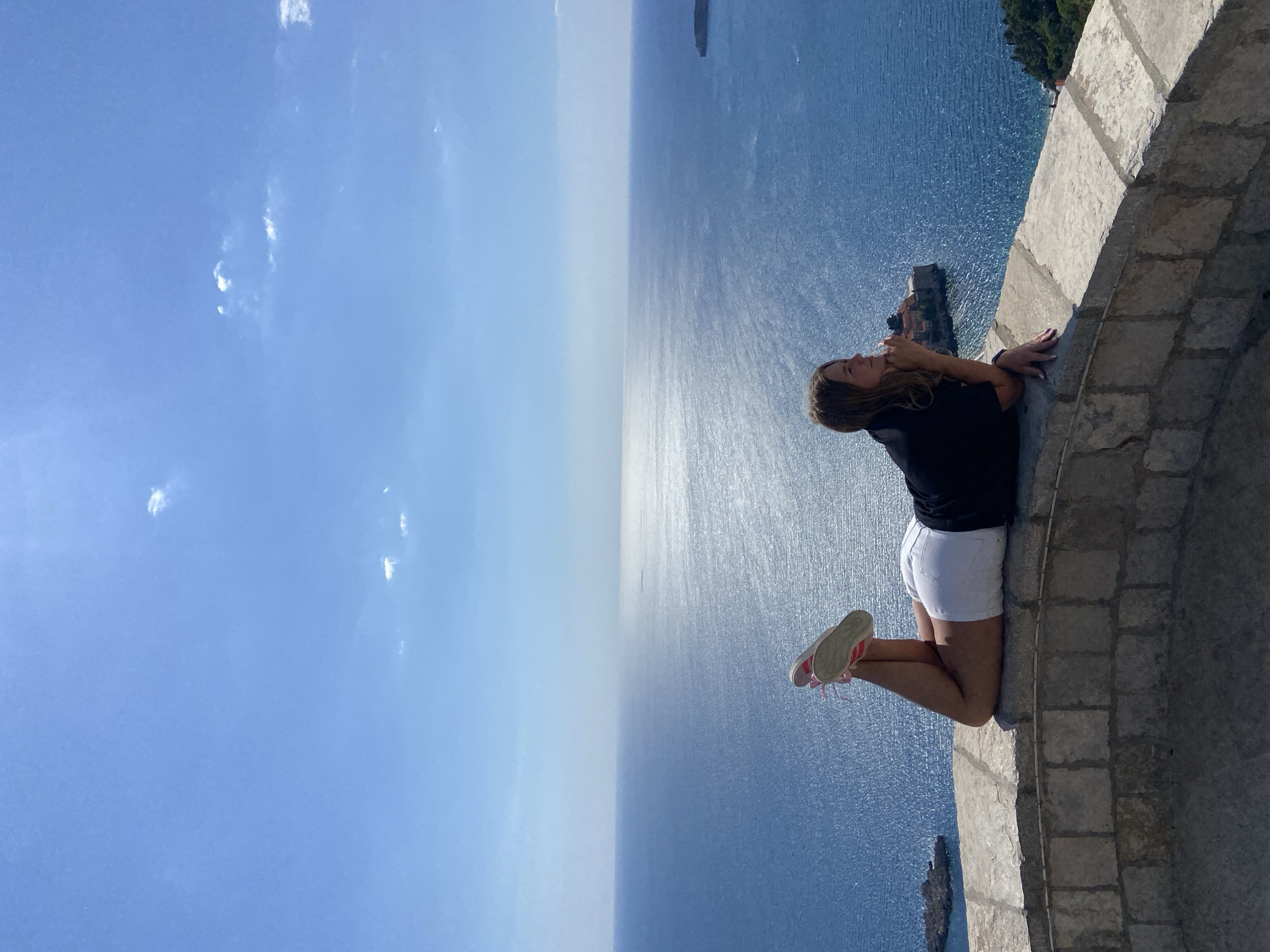 The image depicts a woman kneeling on a stone ledge overlooking the sea in Sveti Stefan, Montenegro. She is wearing a black shirt, white shorts, and red sneakers. She is resting her arms on the ledge and looking out at the water. The sky is blue with a few clouds, and the sea is a deep blue with sunlight reflecting off of it. A small island with buildings on it is visible in the distance. The stone ledge appears to be part of a building or wall.