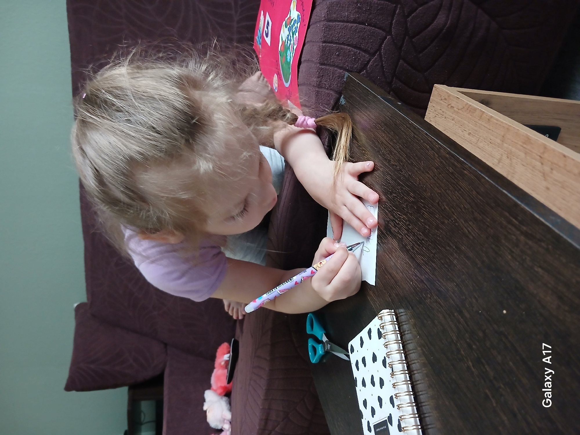 A young child, likely a girl with light-colored hair styled in braided pigtails secured with pink elastic bands, is depicted drawing or writing on a small piece of white paper. She wears a light purple short-sleeved shirt and is barefoot. Her head is tilted, indicating concentration on her task.
The child is seated at a dark brown, possibly wooden, table. In her right hand, she holds a colorful pen or marker with a purple, white, and multi-patterned barrel. To her left on the table are a pair of teal-handled scissors and a spiral-bound notebook with a white cover adorned with black heart or cloud shapes and a gold spiral binding. A red piece of paper or cardboard with an illustration of flowers in a pot and a partially visible number "S" or "5" is positioned near the child's left arm, resting against the back of a brown upholstered couch. The couch features a textured or patterned fabric. The background includes a light green wall and what appears to be a light wooden frame or structure.
The scene suggests a creative play or craft session taking place indoors, possibly in a living room. The photograph includes a "Galaxy A17" watermark in the lower right corner. The activity is occurring in Yekaterinburg, Russia.