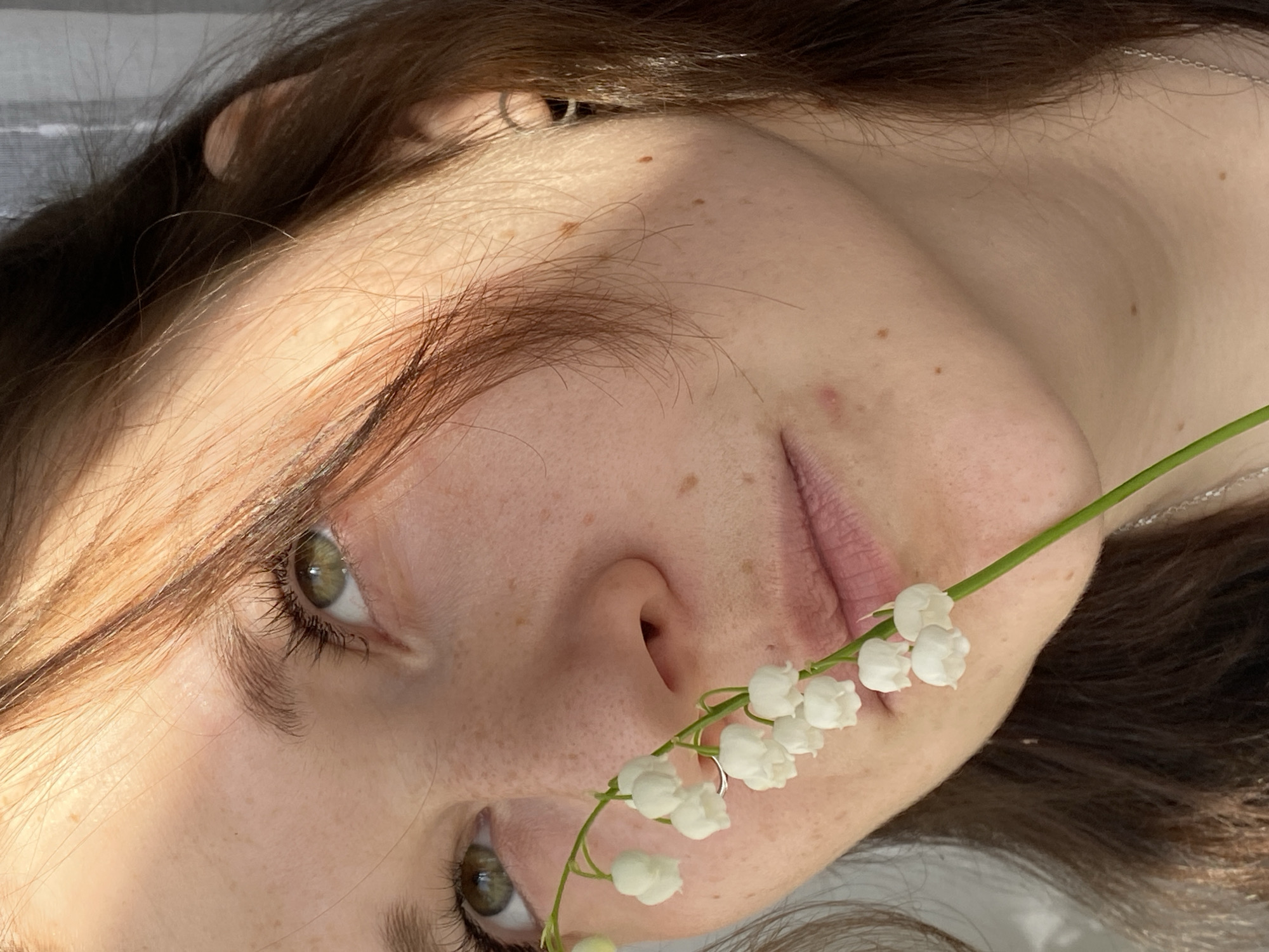 The image features a person, likely a young woman, lying on a white surface. Her face is the main focus, with brown hair framing it. She has light skin with freckles and green eyes. A stem of white lily of the valley flowers is held near her mouth. The lighting is bright, with shadows creating contrast on her face. A silver earring is visible in her ear. The picture does not contain enough information to confirm the location in the city of Chaikovsky, Russia.