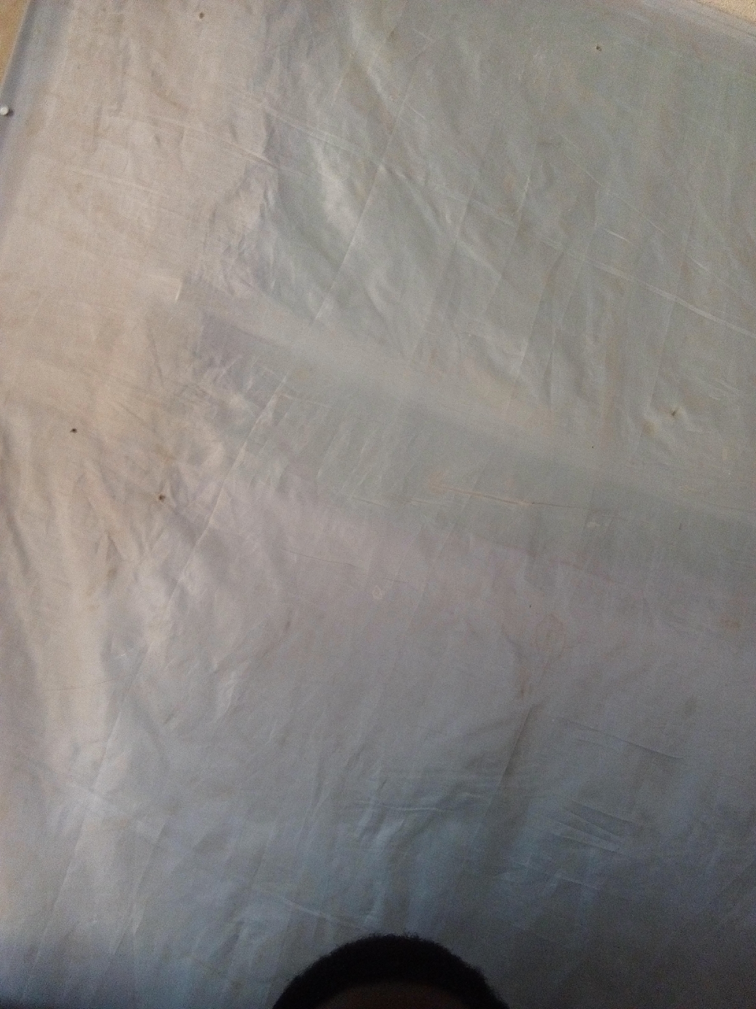 A close-up, low-angle shot captures a wrinkled, light-colored tarp in Nyabe, Nigeria. The tarp appears to be made of a thin, translucent material, possibly plastic. Subtle diagonal lines, indicative of folds or seams, are visible across its surface. There are several small, dark specks scattered on the tarp, suggesting dirt or debris. In the lower right corner, a portion of a person's dark hair and forehead is visible, indicating someone is looking over the tarp from below. The lighting is soft and diffused, creating subtle shadows that highlight the tarp's texture and imperfections. The overall impression is of a simple, utilitarian object with signs of wear and tear.