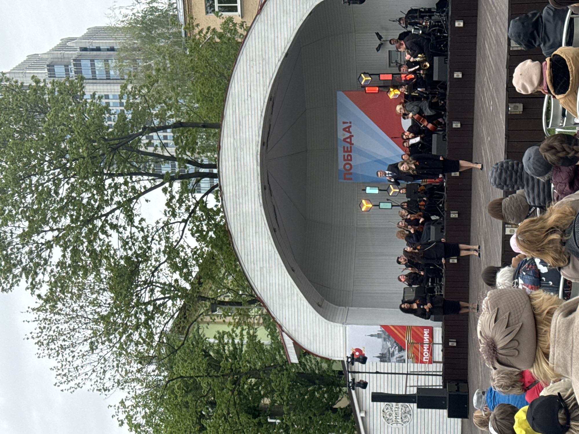 The image shows an outdoor stage with a curved white roof in Moscow, Russia. A small orchestra is performing on stage. A banner with the word "ПОБЕДА!" ("Victory!") in Russian is visible in the background. Spectators are watching the performance from below. A banner on the lower left of the stage reads "ПОМНИМ!" ("We remember!"). A building and trees are in the background.