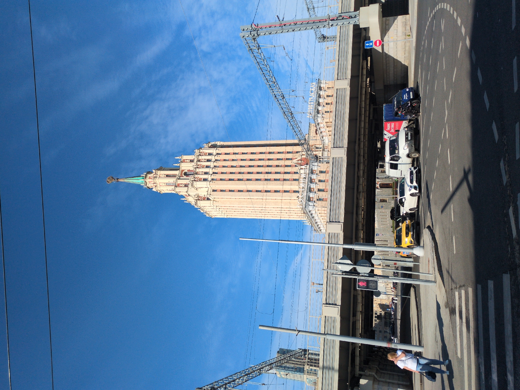 A bright, sunny day in Moscow, Russia, with a clear blue sky and a few wispy clouds. The iconic Stalinist skyscraper, the Hotel Ukraina, stands tall in the background, its distinctive star-topped spire reaching towards the sky. A multi-lane highway overpass runs across the mid-ground, with intricate railway tracks and overhead electrical wires crossing above.
In the foreground, a pedestrian crossing marks the intersection of a road. Several vehicles are visible: a bright yellow car, a white van, a blue delivery truck, and a black motorcycle. A woman in casual clothing, carrying a dark bag, walks on the sidewalk, partially obscured by a traffic light pole. The scene is framed by the shadows cast by the overpass, indicating the sun's position. The overall impression is one of urban bustle under a clear sky.