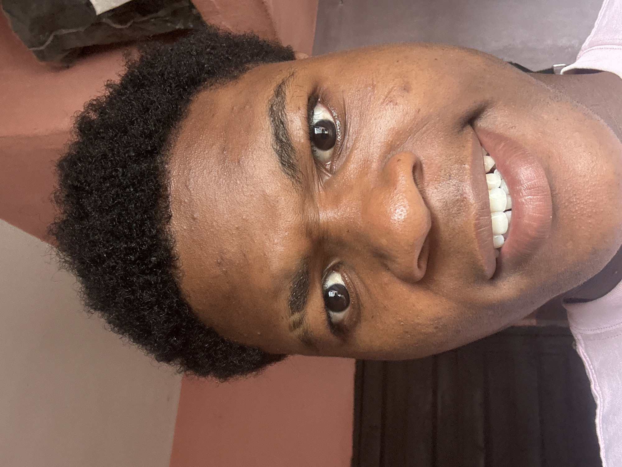 A close-up portrait of a young person, rotated 90 degrees clockwise, captures them smiling broadly and looking directly at the viewer. The person has medium-dark skin, which appears smooth and healthy, and short, tightly curled black hair. Their wide smile reveals a full set of white, well-maintained teeth. Dark brown eyes show distinct reflections of an indoor environment, including a rectangular light source and what appears to be a room interior. The eyebrows are dark and naturally shaped. A light-colored, possibly pink or lavender, t-shirt is visible on the right side of the frame. The background suggests an indoor setting, with a plain wall painted a reddish-brown or terracotta color, transitioning to a darker, possibly wooden or dark-painted surface on the lower right. The image conveys a sense of happiness and warmth from the individual, located in Ihiagwa, Nigeria. No visible text is present.