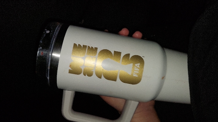 A close-up image depicts a white, insulated travel mug with a light grey handle, held by a human hand. The mug features a prominent gold-colored logo on its side, which reads "WE ARE ONE" with the "O" or "0" stylized as a FIFA World Cup trophy silhouette, followed by "FIFA" in smaller text below. The mug's lid is silver-colored with a clear plastic top, exhibiting visible splashes or residue of a white, milky liquid. The background is uniformly dark and indistinct, suggesting an interior environment with low light conditions or a deep focus on the object. A dark, possibly fabric surface, is visible in the lower foreground. No specific location within Tlaxcala City, Mexico, is discernible from the visual evidence provided.