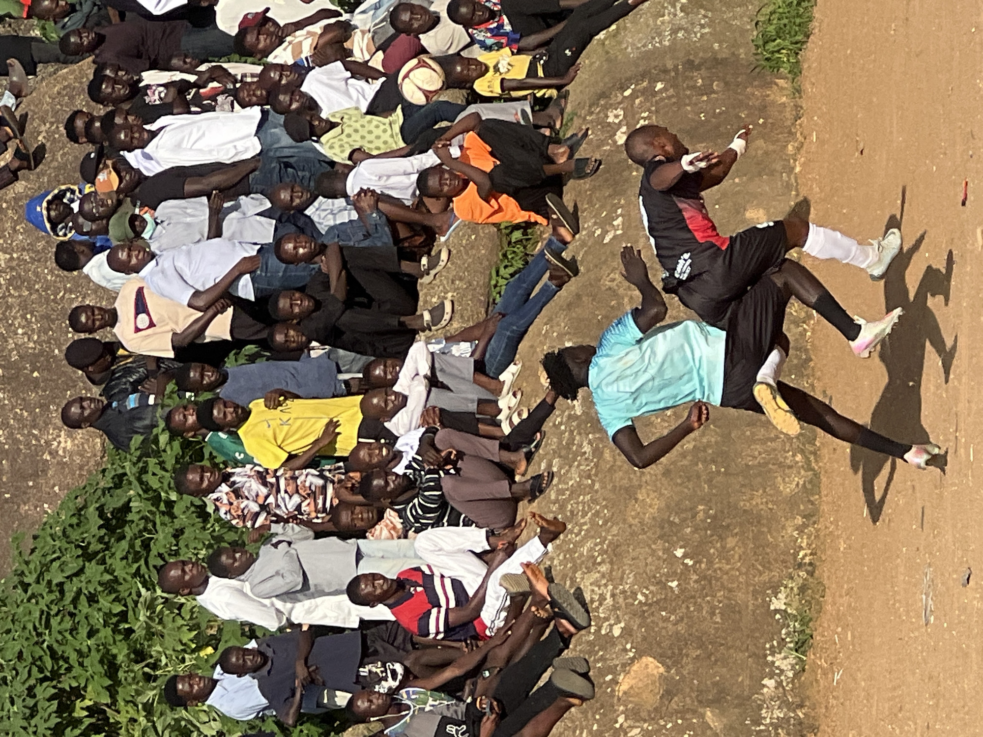 In the city of Jos, Nigeria, a group of men and boys are gathered on a rocky incline, observing an event unfolding on a dirt field below. The scene is dominated by the natural environment, with a large rock formation providing seating for many of the spectators.
The spectators are a mix of ages, with many appearing to be young men. They are dressed in a variety of casual clothing, including t-shirts, shirts, and trousers. Some are seated on the rocks, while others stand behind them.
On the dirt field below, two young men are engaged in a sporting activity, possibly a football match. One is wearing a blue t-shirt and black shorts, while the other is in a black and red jersey with black shorts. They are both in motion, suggesting an active game.
A football is visible in the background, near a group of seated spectators, and the overall atmosphere suggests a casual, community-based sporting event. The background shows dense green foliage and the textured surface of the rocky terrain. The lighting indicates it is daytime, with sunlight casting shadows.