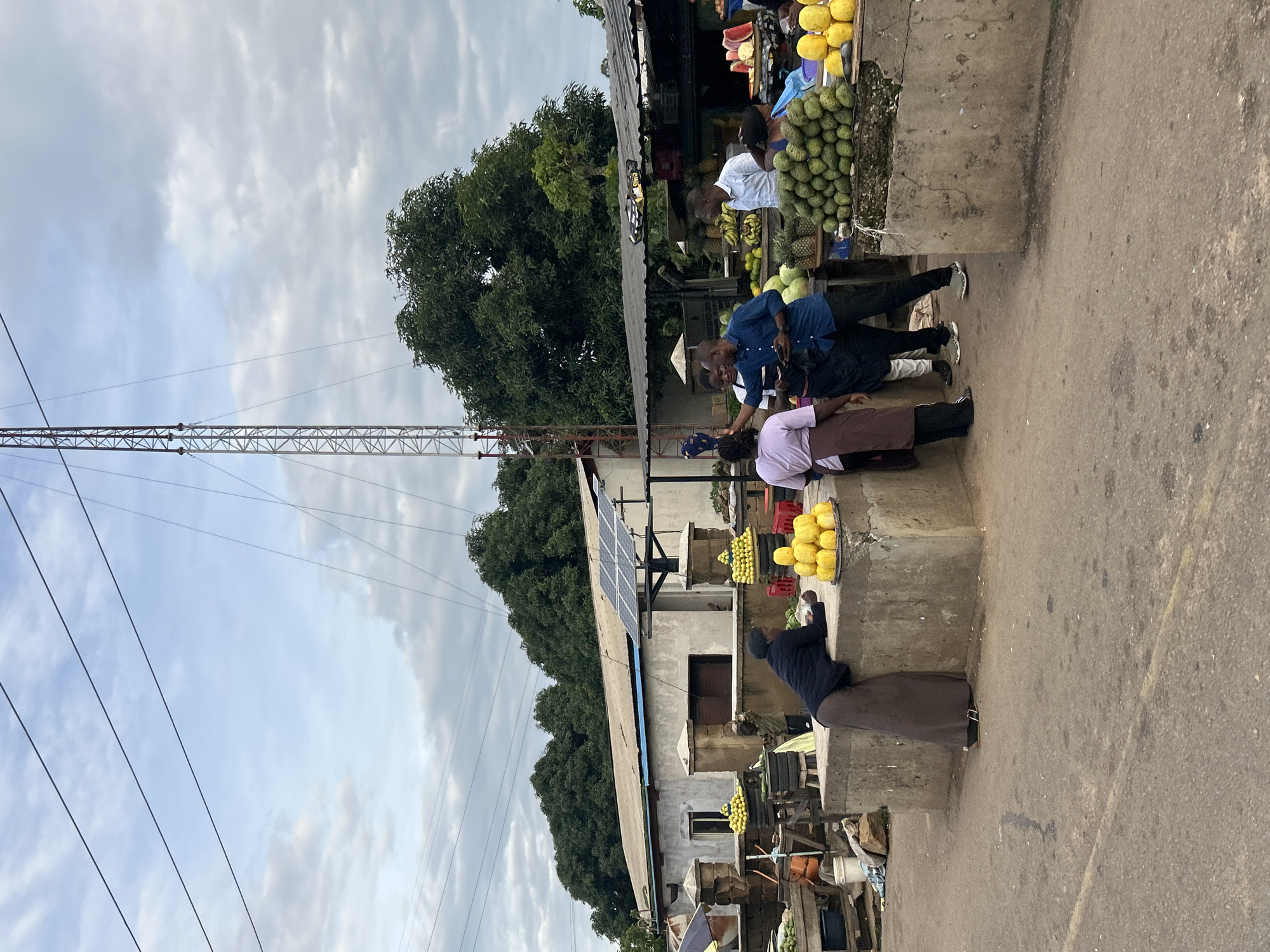 The image depicts a bustling outdoor market scene in Jos, Nigeria, under a cloudy sky. A tall, metal telecommunications tower dominates the background, with power lines extending across the frame. Several stalls display a variety of fruits, including piles of green soursop, yellow mangoes or papayas, pineapples, and bananas.
In the foreground, a woman in a long brown skirt and dark top leans over a concrete market stall, resting her arms. To her right, a group of people are gathered around another stall. A man in a blue shirt and dark pants is holding a mobile phone and gesturing. A woman in a purple top and brown apron stands nearby, and another man in a white short-sleeved shirt is also present.
Behind the stalls, a building with solar panels on its roof is visible, suggesting an attempt at sustainable energy. The ground is paved with asphalt, indicating an urban setting. The overall atmosphere appears lively, with people actively engaged in commerce and interaction within the market environment.