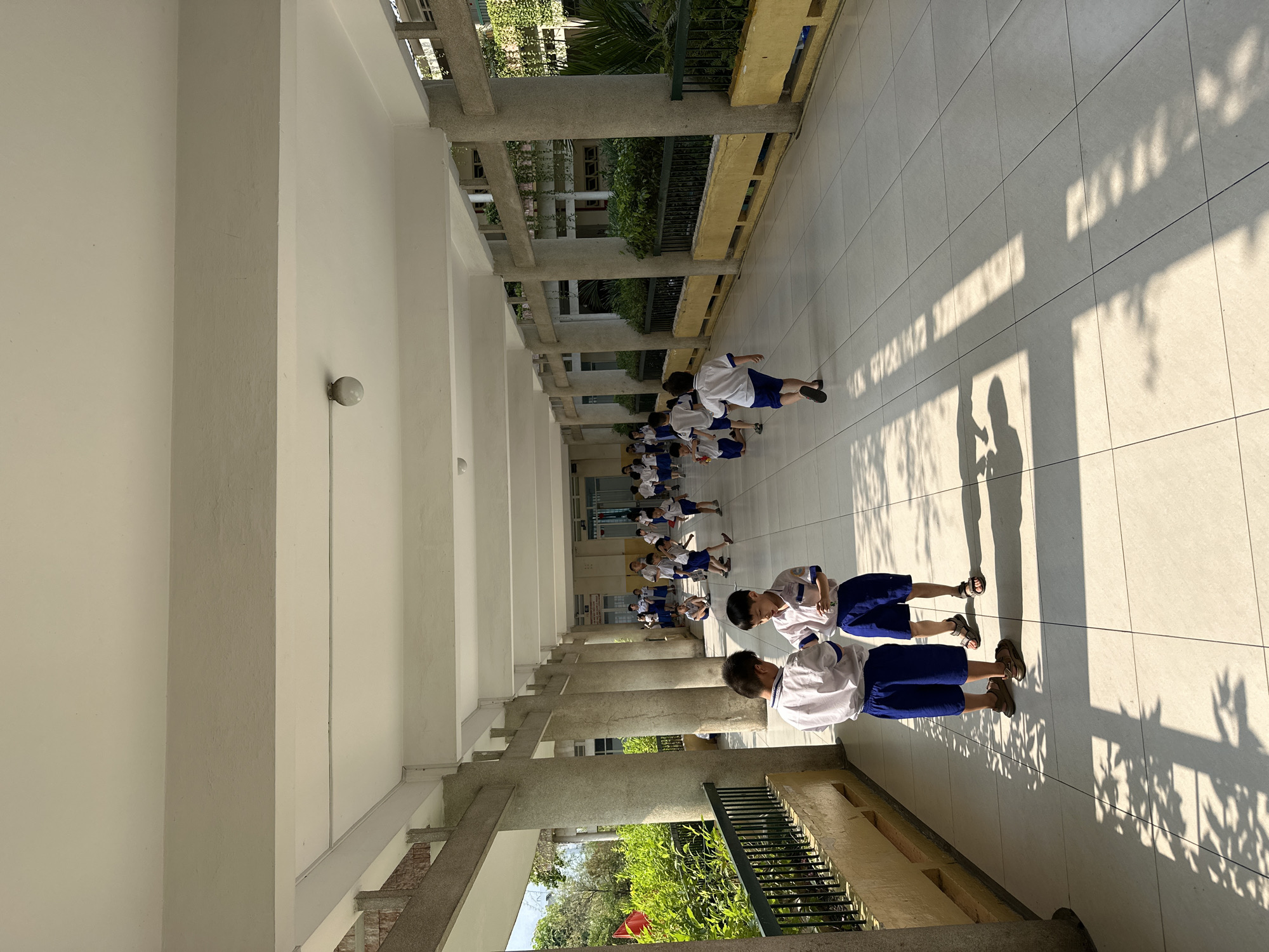 A medium shot depicts a group of children in school uniforms. They are on a bright tiled corridor lined with pillars, likely in Ho Chi Minh City, Vietnam. Some are running, while others are standing. Sunlight filters through nearby trees, casting shadows on the floor.