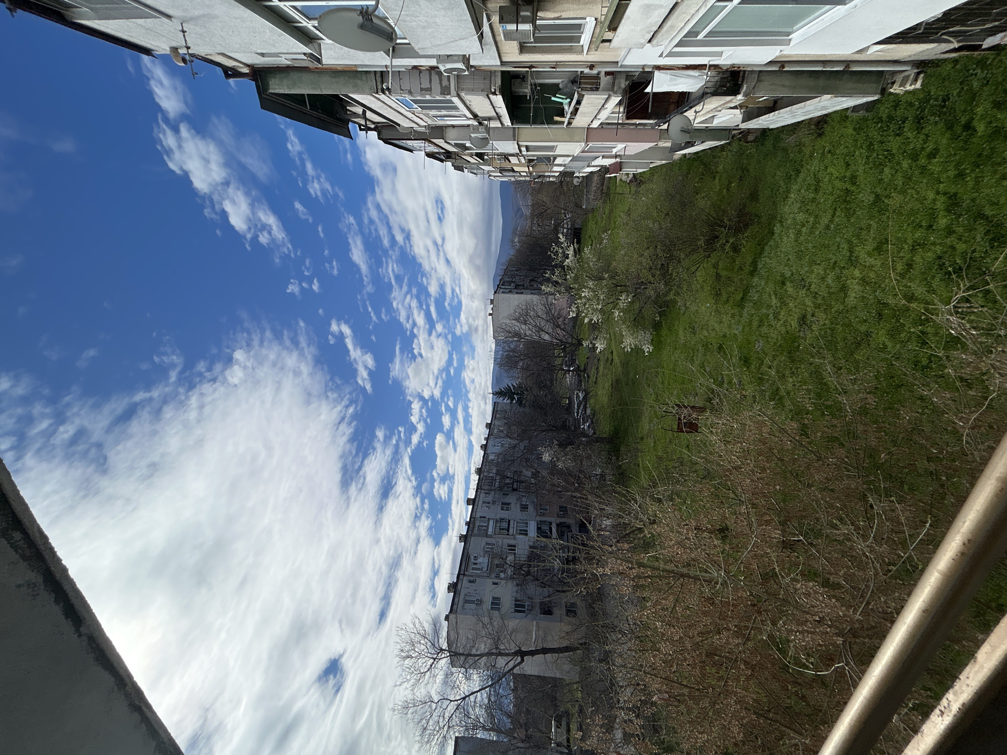 The image depicts a view between two apartment buildings in Plovdiv, Bulgaria. The perspective is from a high vantage point, likely a balcony, looking down into a grassy courtyard or open space. The buildings are typical Eastern European apartment blocks, characterized by a blocky, utilitarian design with visible balconies, satellite dishes, and air conditioning units. The space between the buildings includes grass, bare trees, and trees beginning to bloom, suggesting early spring. A distant mountain range is visible on the horizon. The sky is blue with numerous white clouds.